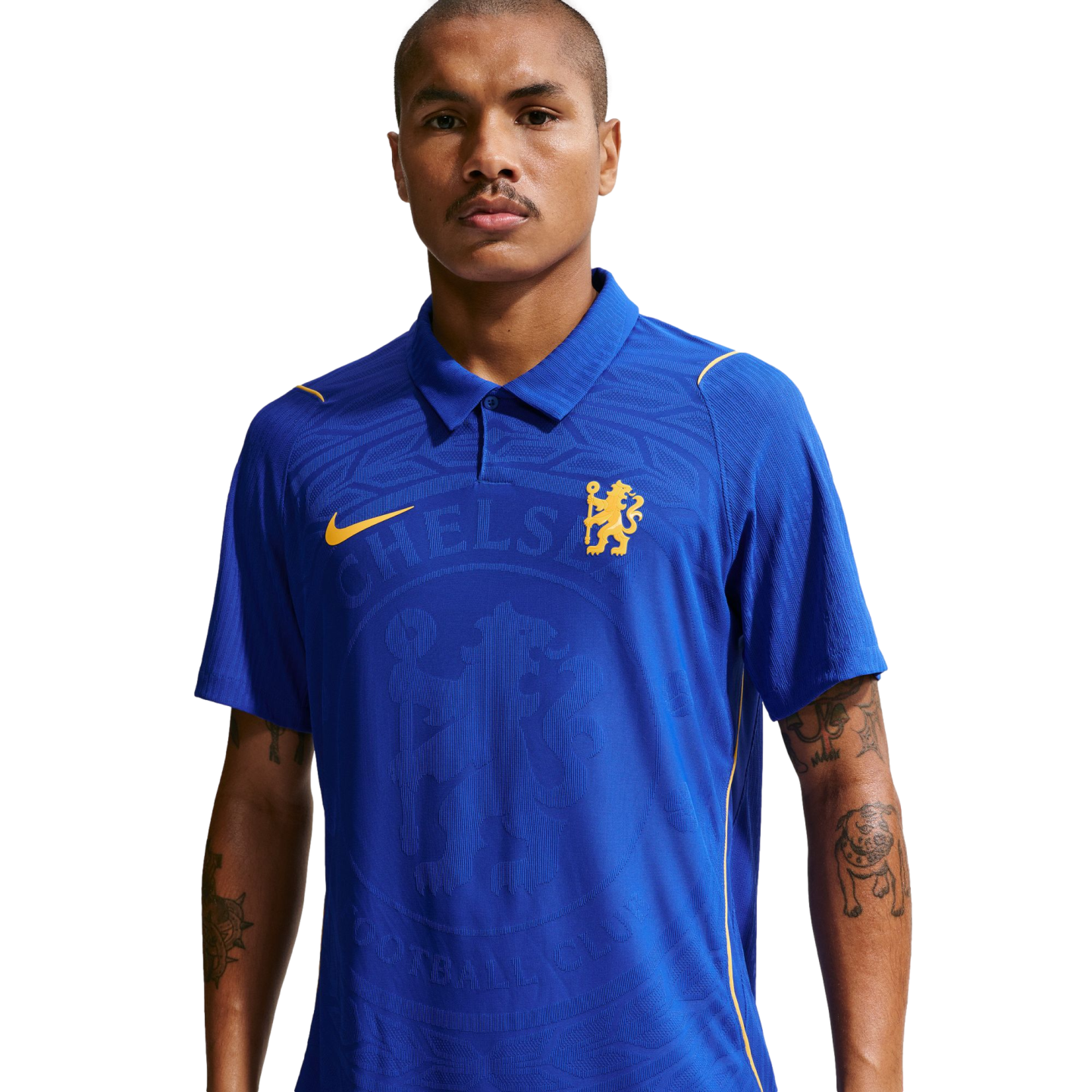 A man wearing a blue, collared Chelsea FC jersey with yellow logos and a large textured crest on the chest, isolated on a white background.