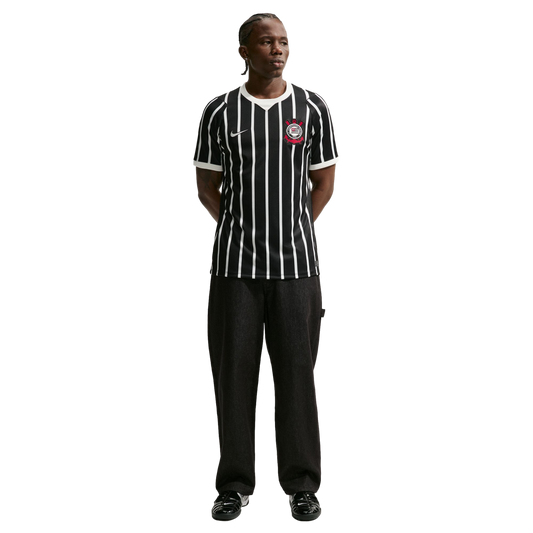 Full-length shot of a man in a black and white striped Corinthians soccer jersey, black pants, and black sneakers, standing against a solid black background.