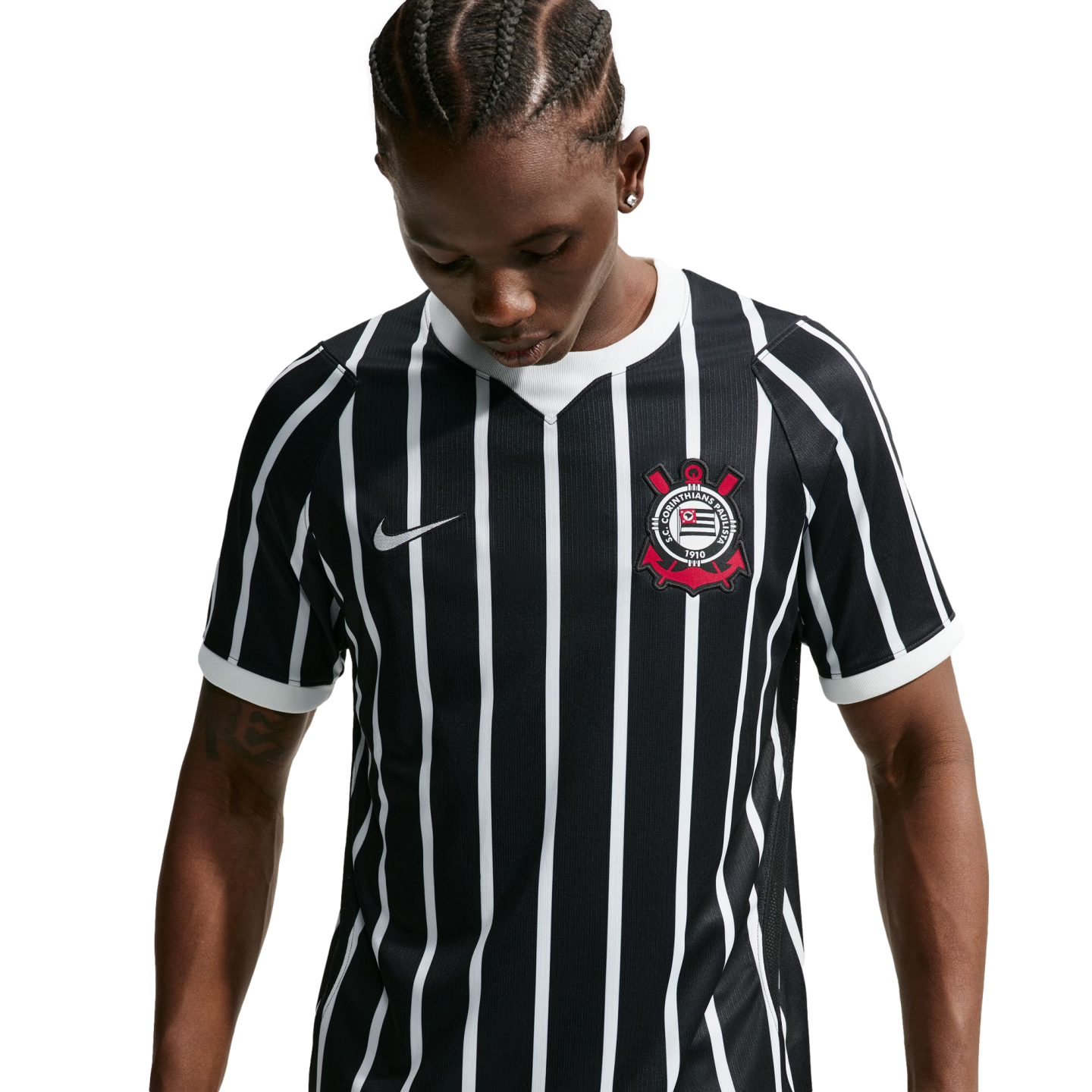 A person with braided hair looks down, wearing a black and white vertically striped Nike soccer jersey with a red and white crest. The background is solid black.