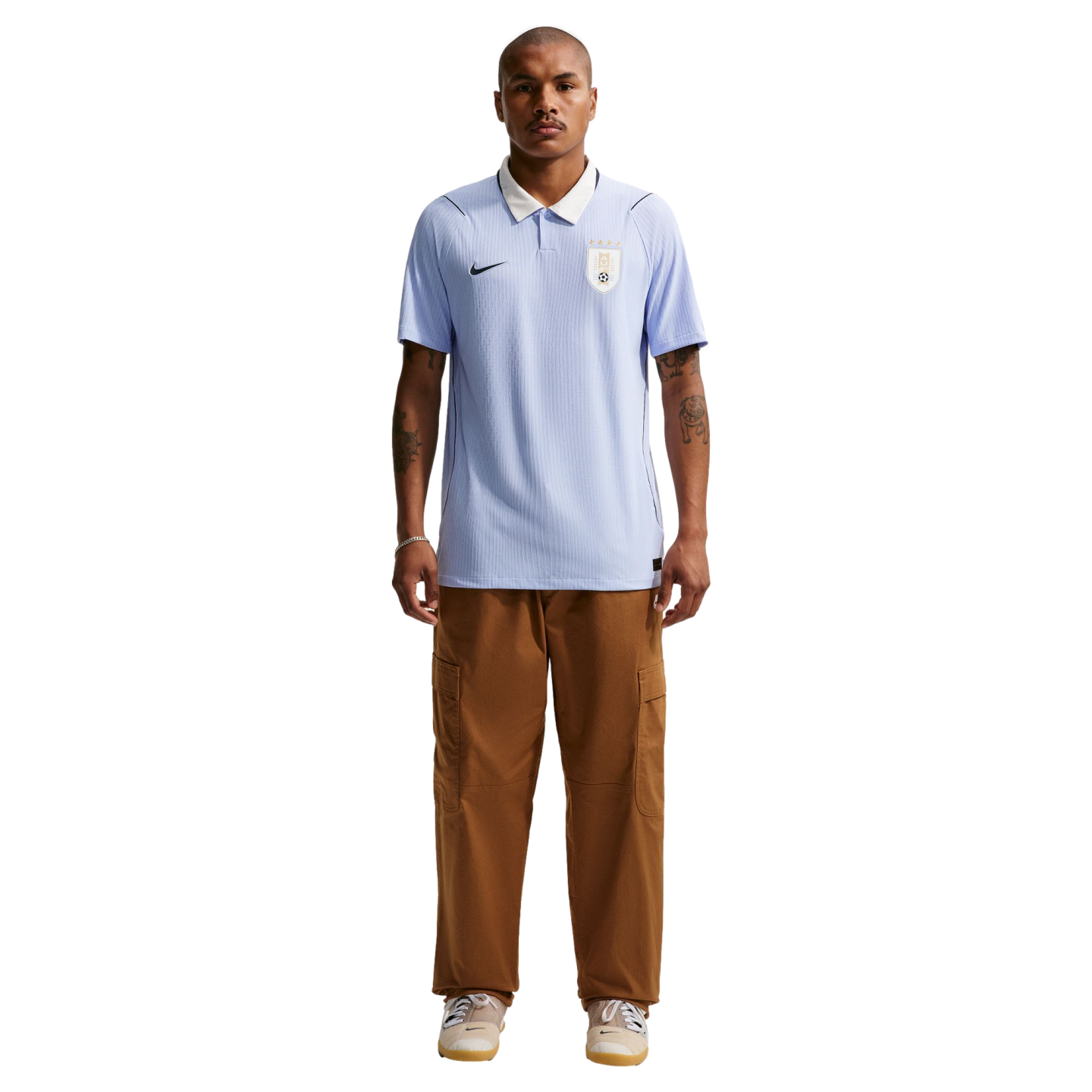 Full-body portrait of a man wearing a light blue Uruguay national team soccer jersey with a white collar, paired with brown cargo pants and beige sneakers against a solid black background.