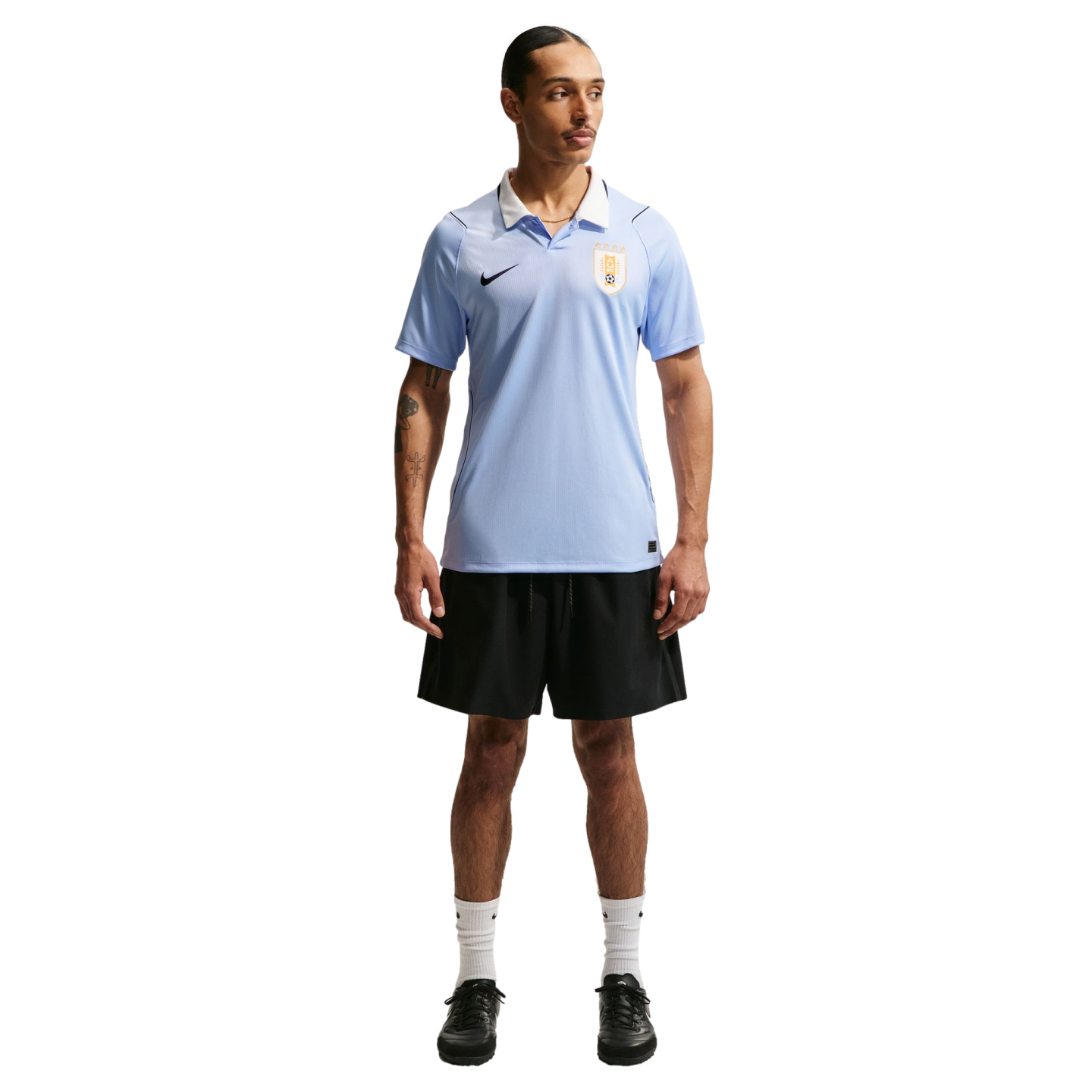 A man wearing a light blue Uruguay soccer jersey, black shorts, and white socks, standing against a black background.