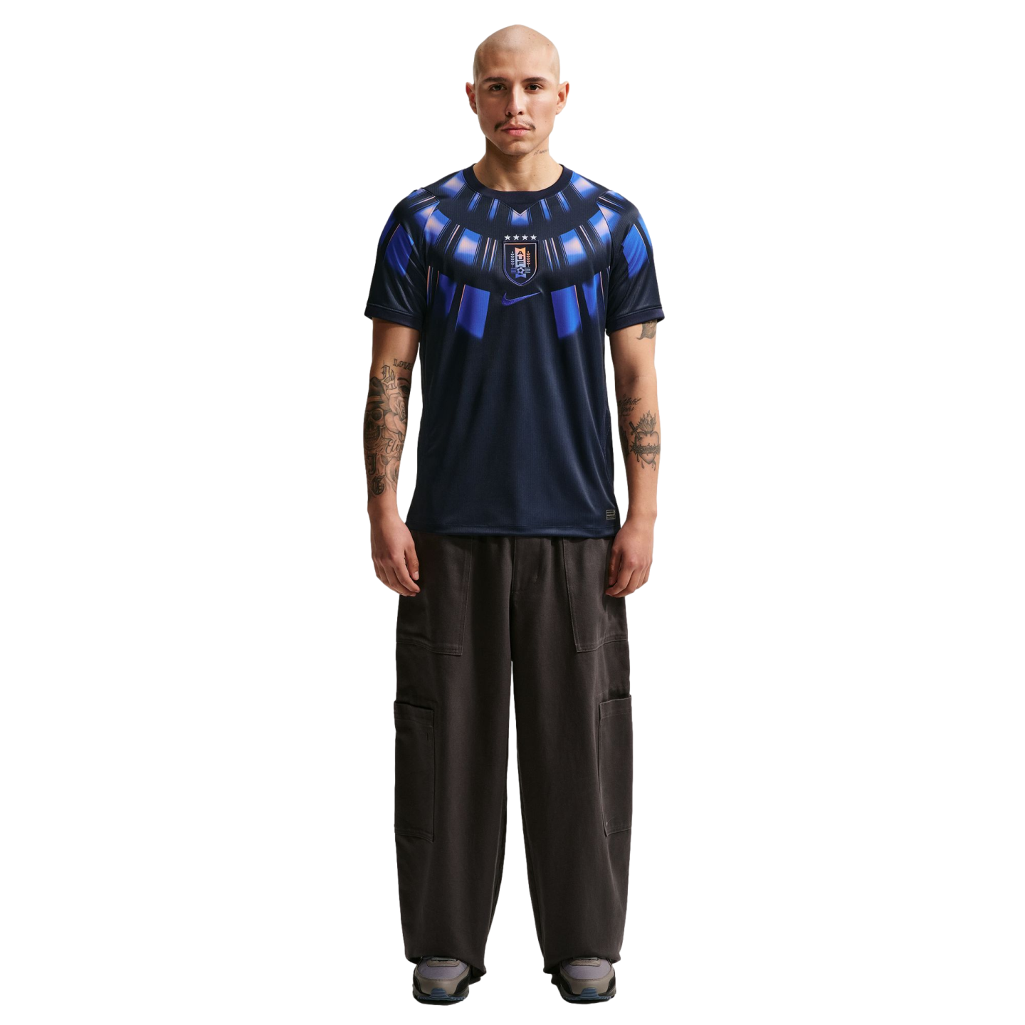 A full-body studio portrait of a bald man with arm tattoos standing against a solid black background. He is wearing a dark navy blue Nike t-shirt featuring a bright blue radial graphic around the collar and a center-chest Uruguay national team crest. His 
