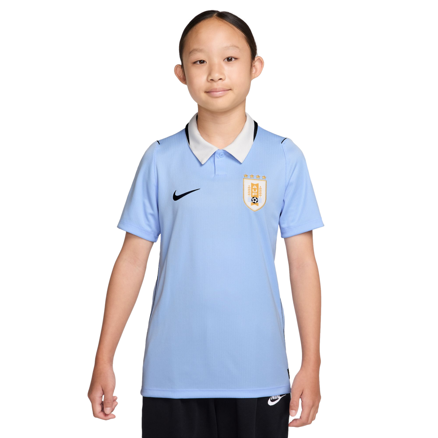 A young girl wears a light blue Nike Uruguay soccer jersey with a white collar and black shorts against a solid black background.