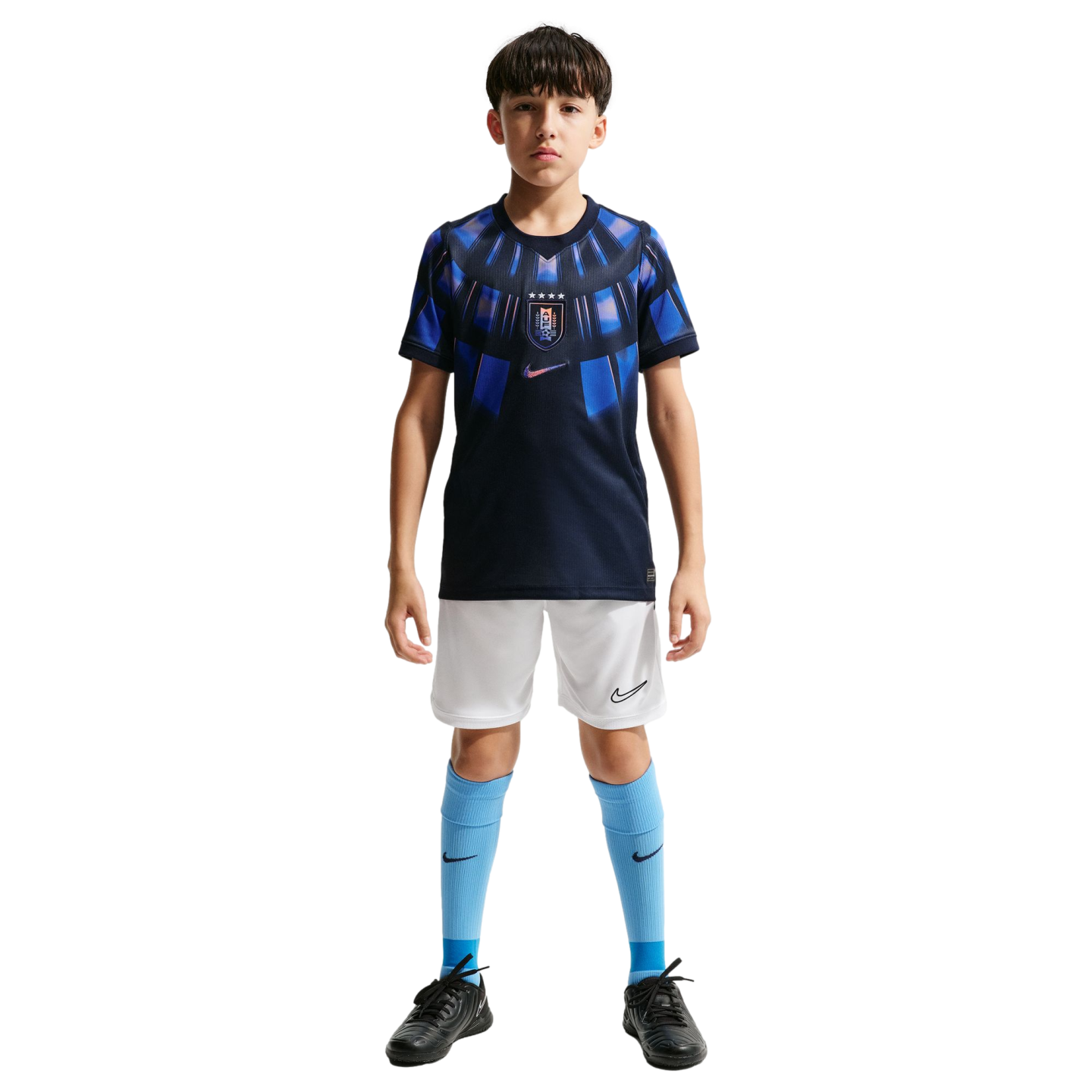 A young boy stands against a solid black background wearing a navy and light blue Nike soccer kit, white shorts, light blue socks, and black soccer cleats.