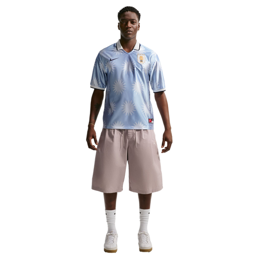 A man wearing a light blue sun-patterned soccer jersey, tan shorts, and white sneakers stands against a plain white background.
