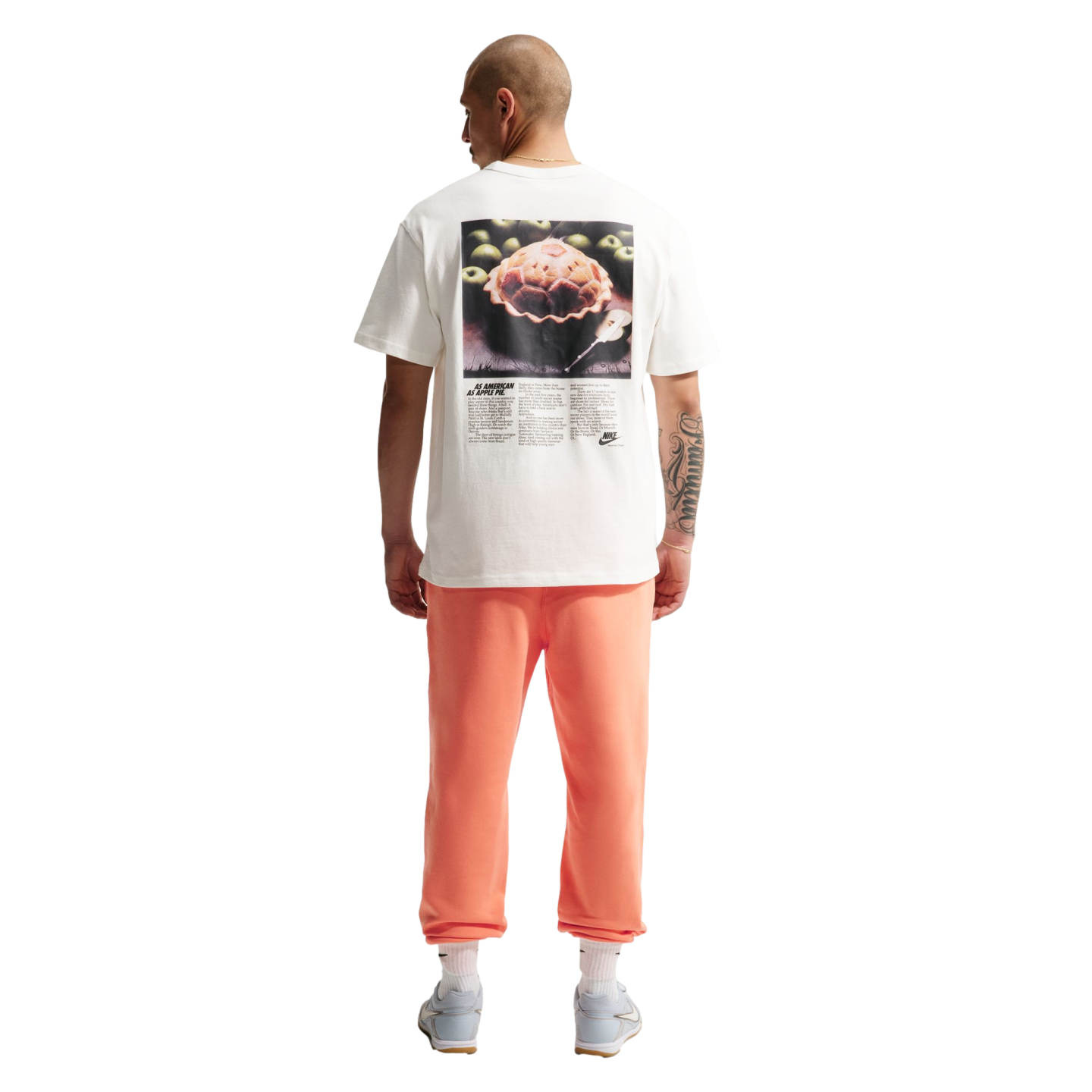 A back view of a man standing against a black background, wearing a white t-shirt with a large graphic of an apple pie and the text "AS AMERICAN AS APPLE PIE." He is also wearing coral-colored sweatpants, white socks, and light blue sneakers. He has a bal