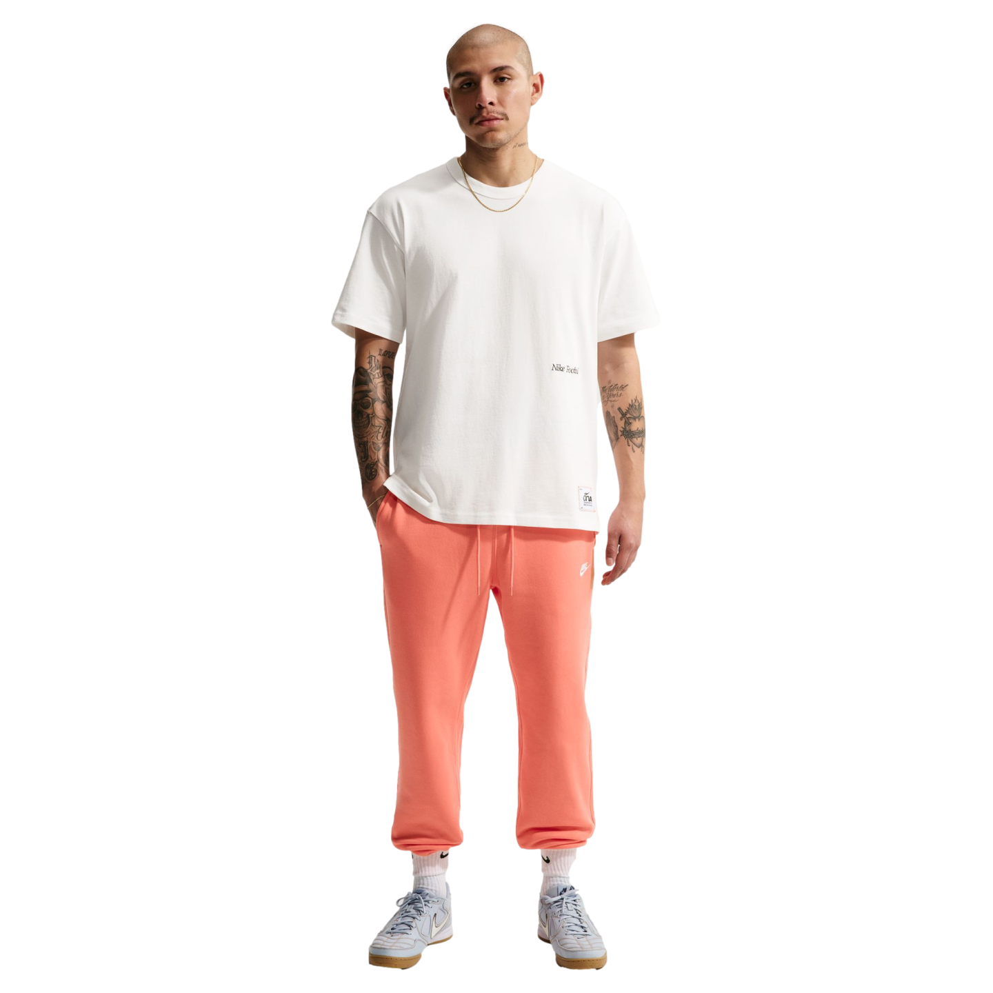 Full-length photo of a bald man with arm tattoos standing against a black background, wearing a white T-shirt, salmon-colored sweatpants, and light blue sneakers.