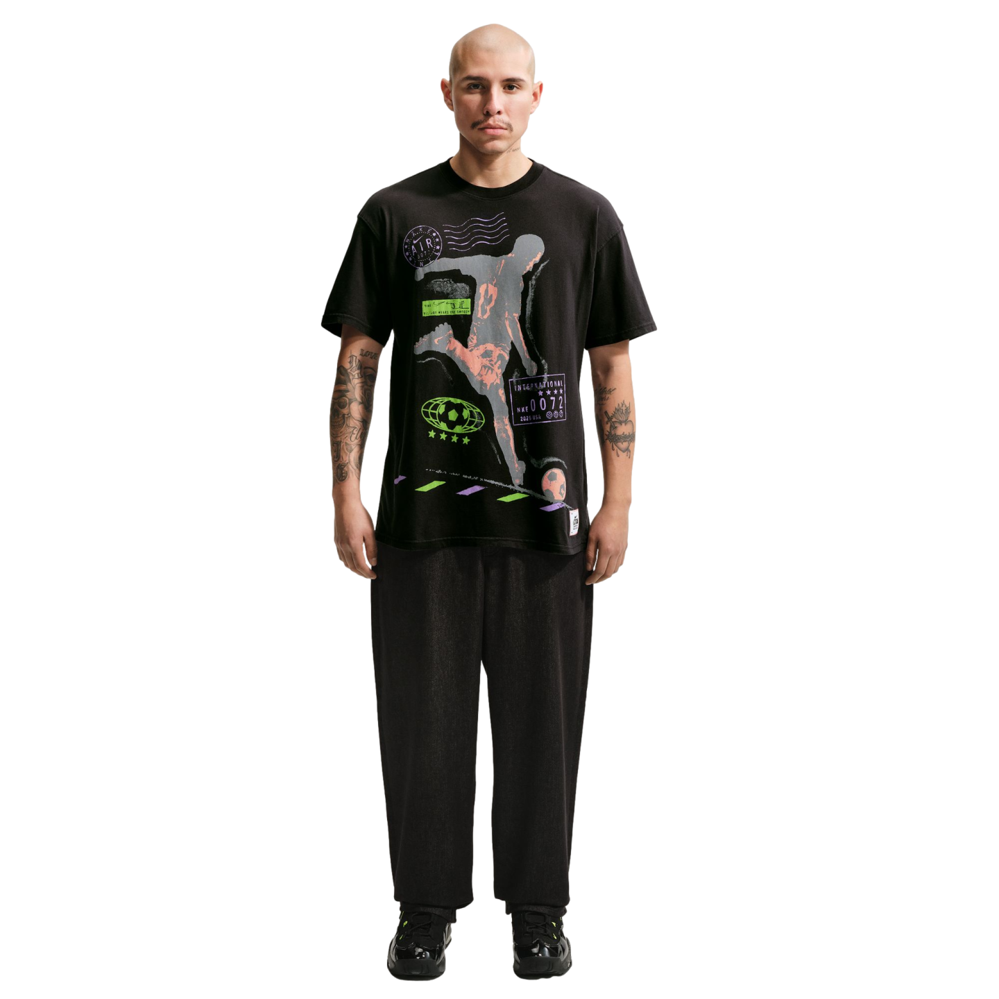 A full-body portrait of a bald man with a mustache and arm tattoos, standing against a solid black background. He is wearing a black graphic t-shirt with a soccer player illustration, loose black trousers, and black sneakers with neon green details.