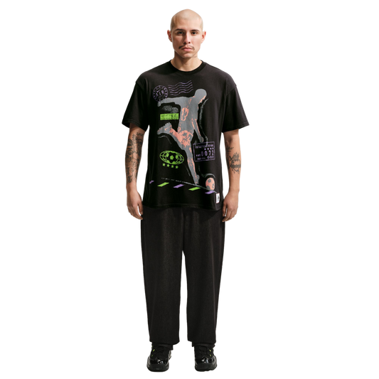 A full-body portrait of a bald man with a mustache and arm tattoos, standing against a solid black background. He is wearing a black graphic t-shirt with a soccer player illustration, loose black trousers, and black sneakers with neon green details.