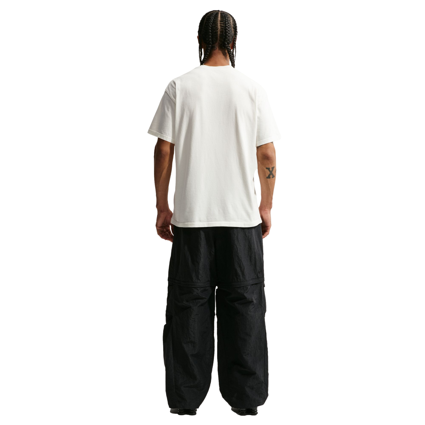 A full-body rear view of a man with braided hair standing against a solid black background. He is wearing a plain white short-sleeved t-shirt and oversized, black technical pants. A small "X" tattoo is visible on his right forearm.