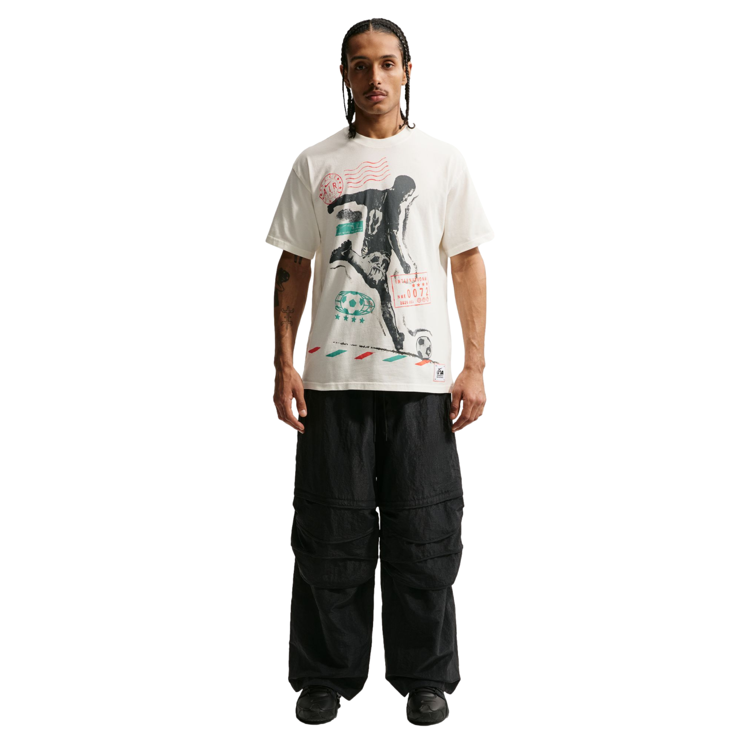 A man stands against a solid black background wearing a white graphic t-shirt featuring a soccer player silhouette, oversized black cargo pants, and chunky black sneakers.