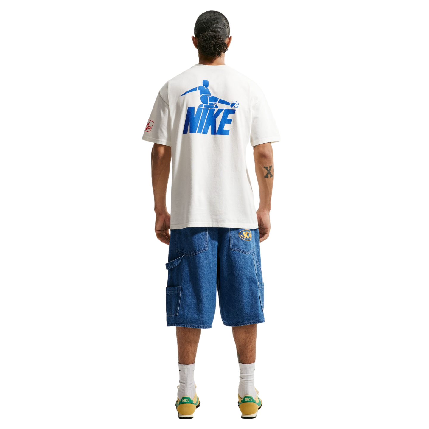 A person stands with their back to the camera against a black background, wearing a white t-shirt featuring a blue Nike soccer graphic on the back, loose-fitting blue denim shorts, white crew socks, and yellow and green sneakers.