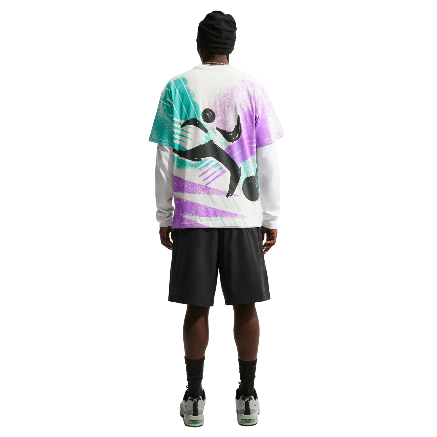 A back view of a person standing against a solid black background, wearing a black beanie, a colorful graphic t-shirt layered over a white long-sleeved shirt, black shorts, and grey sneakers.