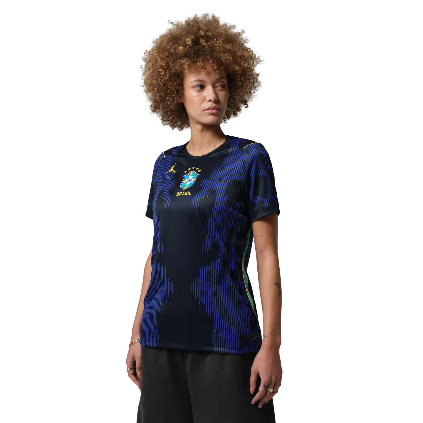 A person with a large curly afro wearing a blue and black patterned Brazil national team jersey, looking to the side against a black background.