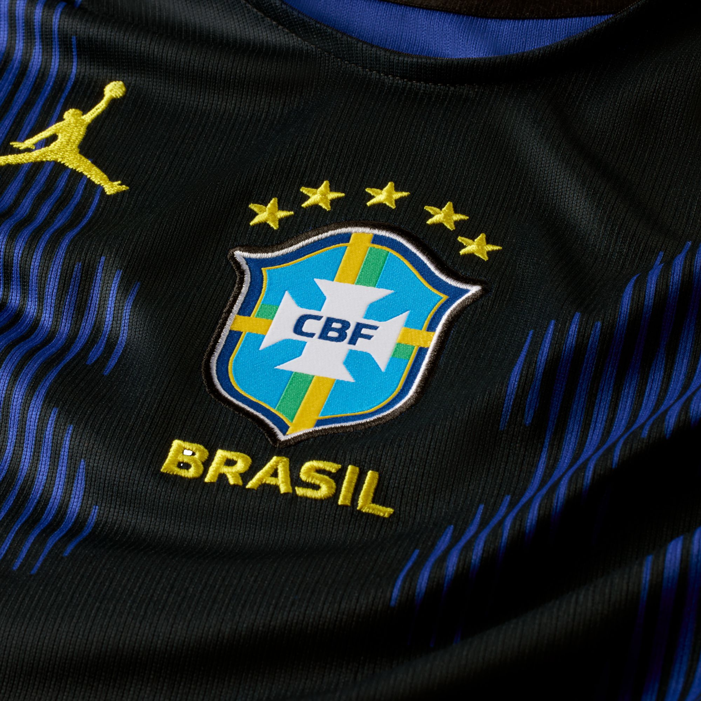 Close-up of a black Brazil soccer jersey featuring the CBF crest, five stars, "BRASIL" text, and a yellow Jumpman logo against a background of blue vertical patterns.