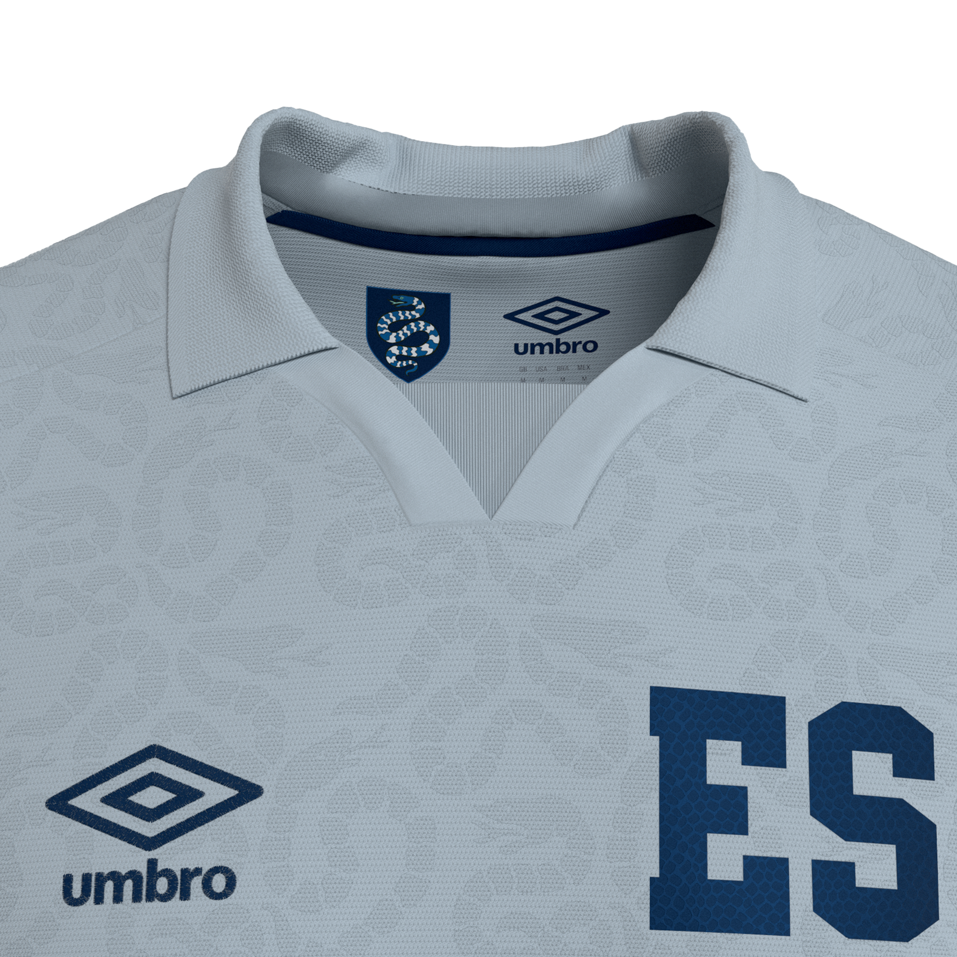 Close-up of a light blue Umbro polo jersey with a large "ES" on the chest and a snake emblem inside the collar. The fabric features a subtle, all-over textured pattern.