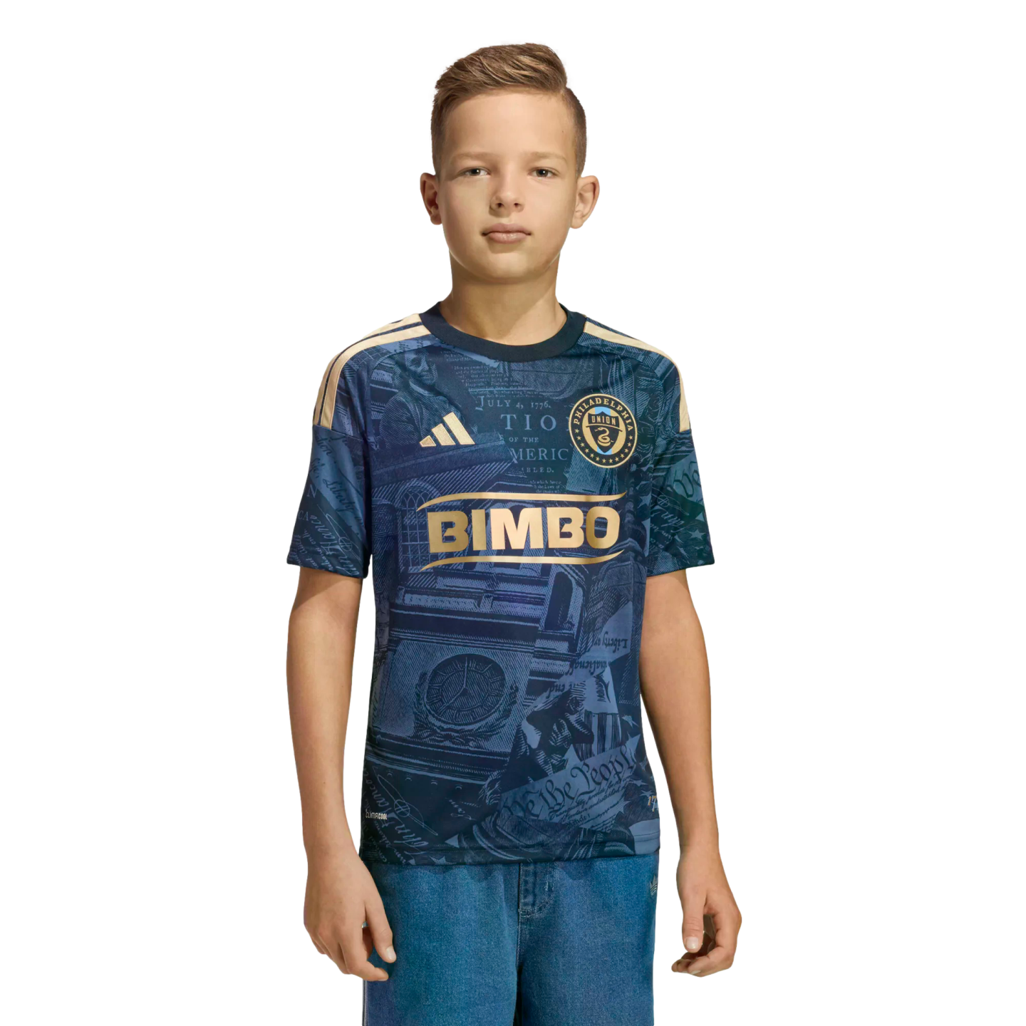 A youth stands wearing a navy blue and gold Philadelphia Union soccer jersey and blue jeans against a solid black background.