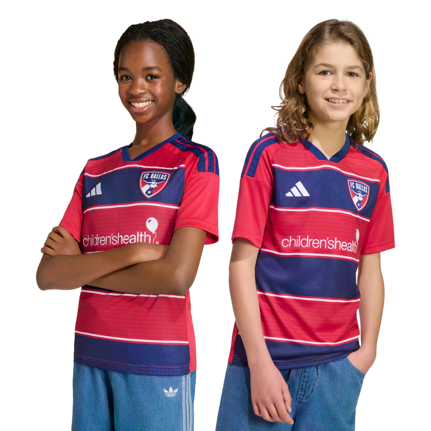 Two smiling young fans wearing red and blue striped FC Dallas home jerseys and blue shorts, posing against a black background.