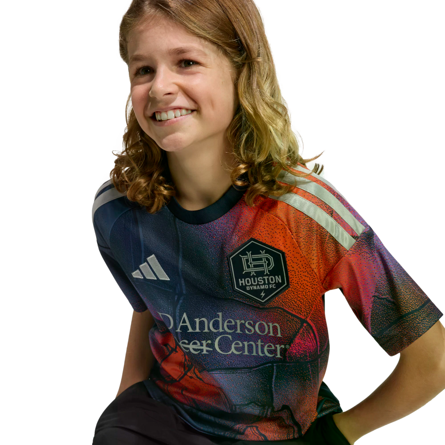 A smiling child with long, wavy light brown hair wears a colorful Houston Dynamo FC soccer jersey against a solid black background.