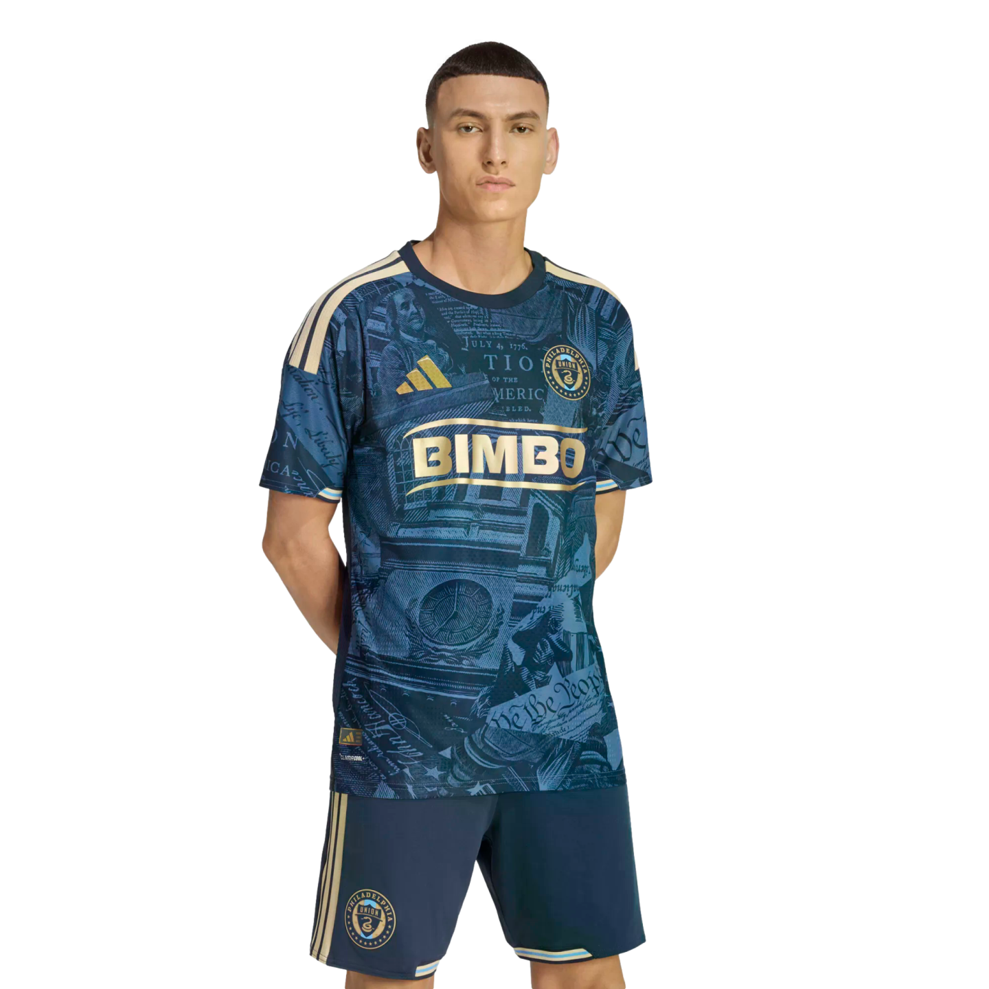 A man models a navy blue and gold Philadelphia Union soccer kit featuring historic Philadelphia-themed graphics, standing against a black background.