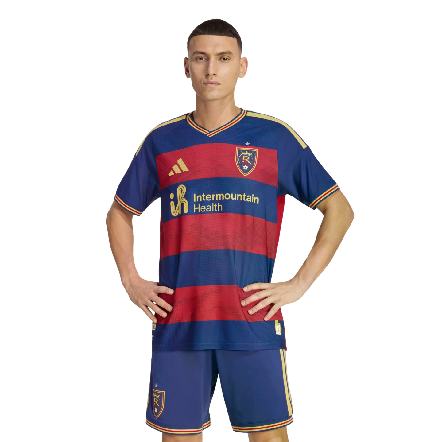A man models a blue and red striped Real Salt Lake soccer jersey and blue shorts. He stands with hands on his hips against a black background.