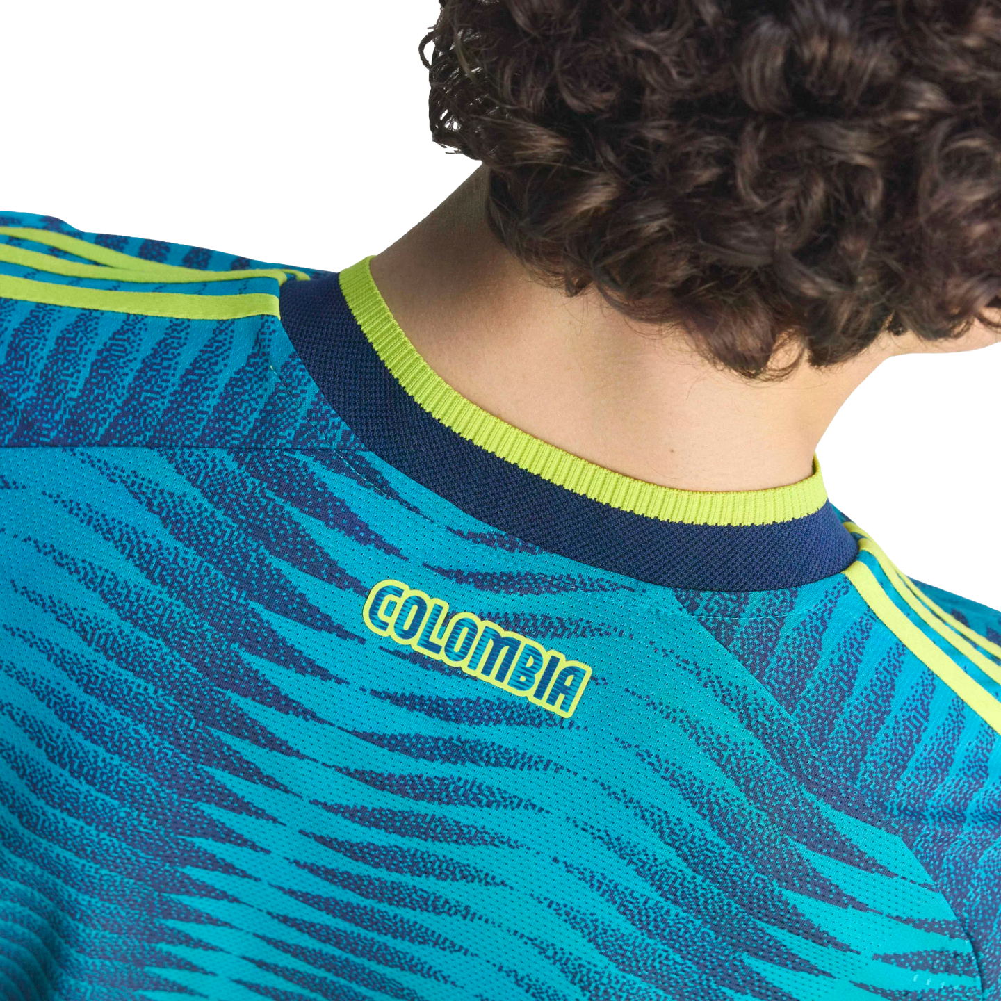 Back view of a person with curly hair wearing a teal patterned soccer jersey with yellow "COLOMBIA" text and lime green accents.
