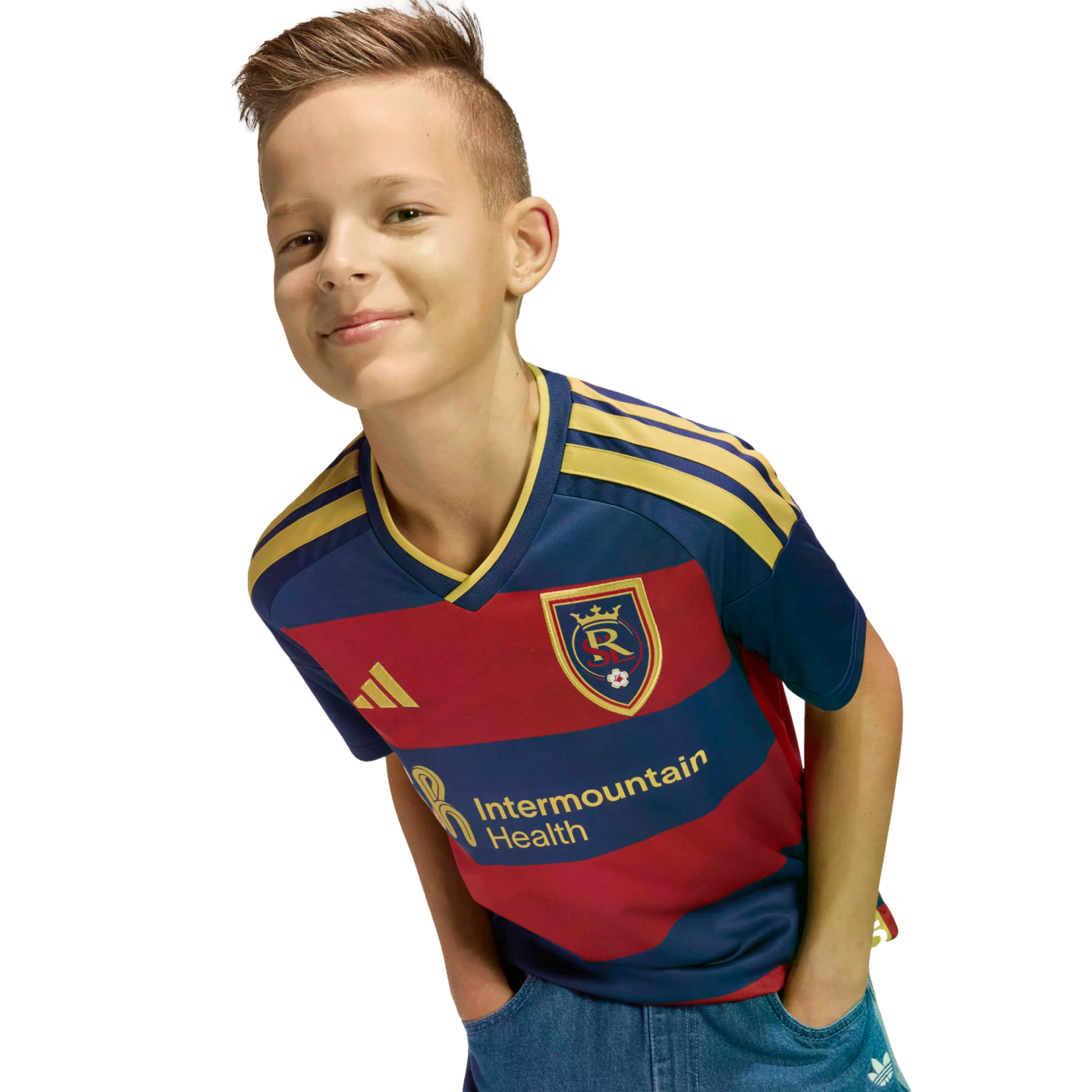 A smiling boy wearing a navy and red Real Salt Lake soccer jersey leans forward against a black background.