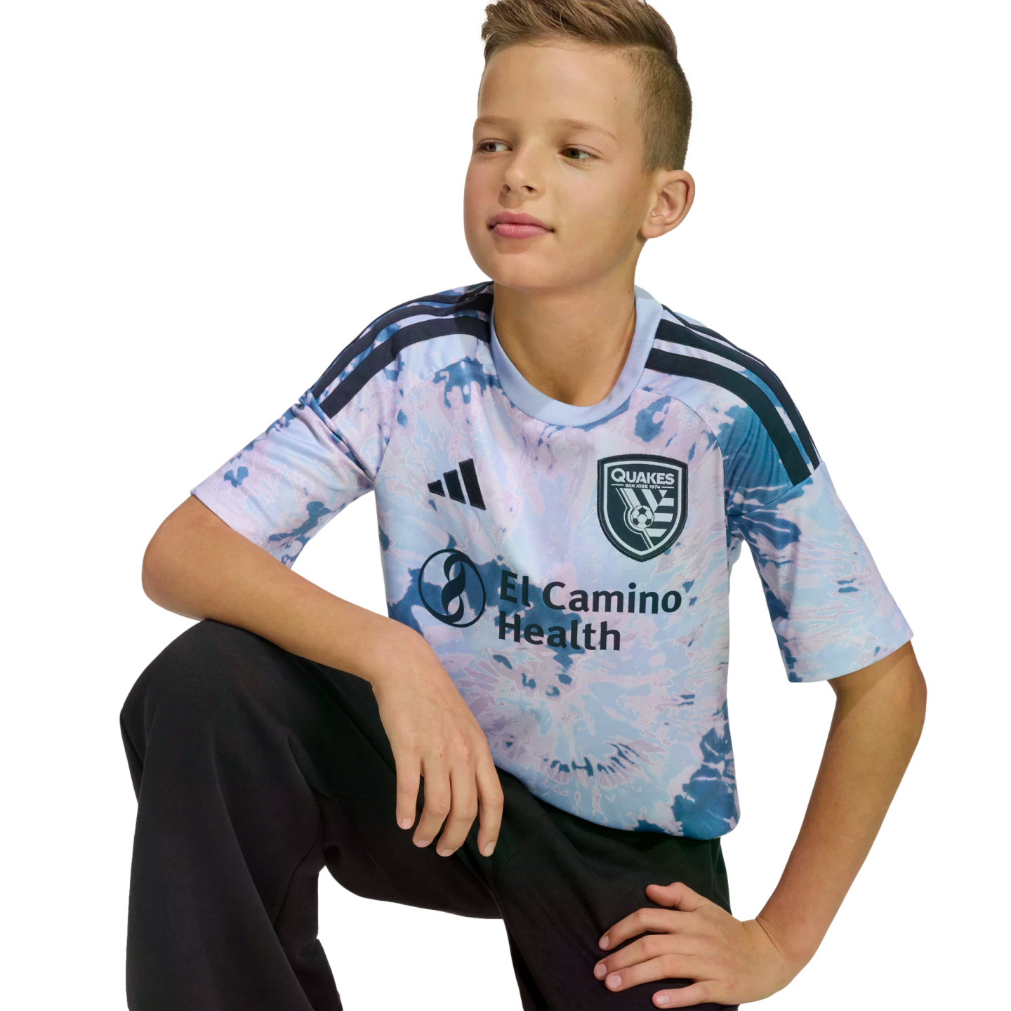 A young boy with brown hair kneeling, wearing a tie-dye San Jose Earthquakes soccer jersey and black pants against a black background.