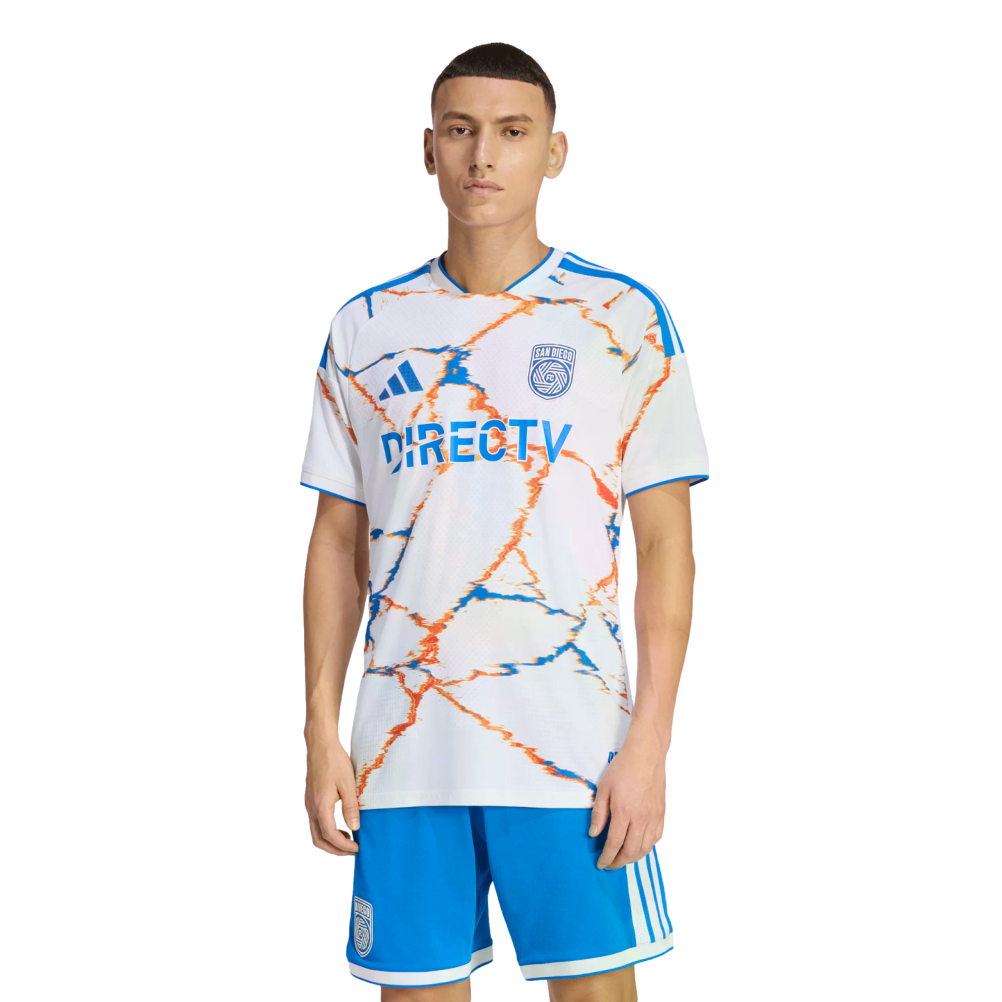 A man models a San Diego FC soccer kit with a white jersey featuring orange and blue abstract patterns and a DIRECTV logo, paired with blue shorts.