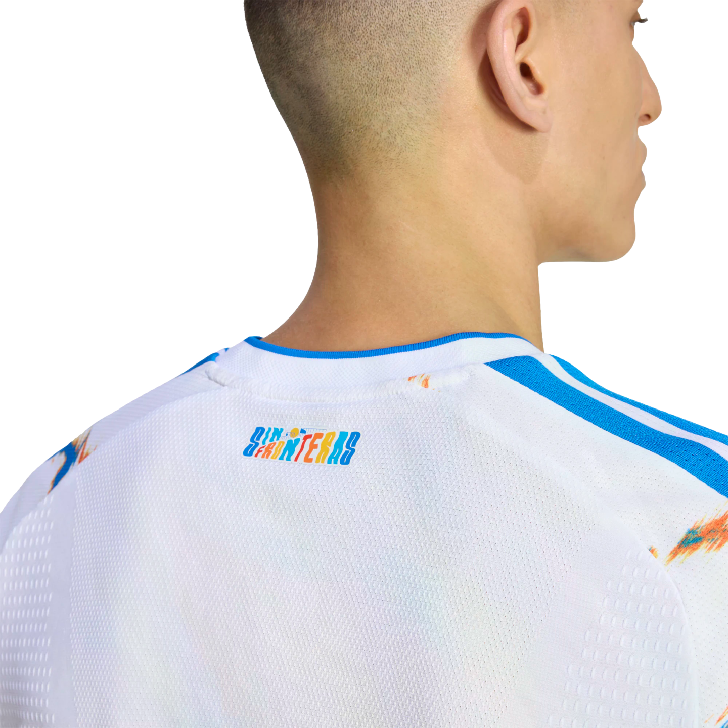 A close-up of the back of a man's neck and upper back, wearing a white jersey with blue trim and a colorful "SIN FRONTERAS" graphic on the neck.