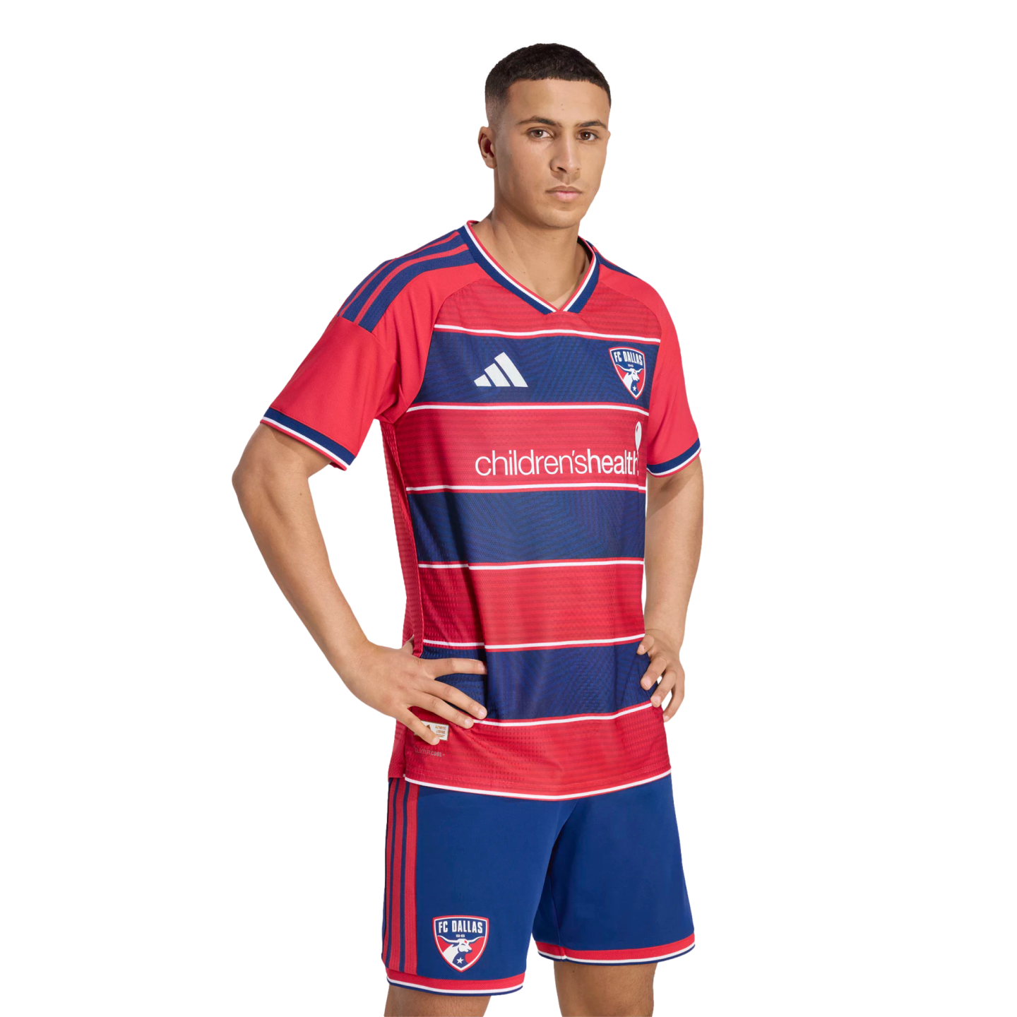 Male model posing in the FC Dallas home soccer kit, featuring a red and blue striped jersey with "children'shealth" logo and matching blue shorts.