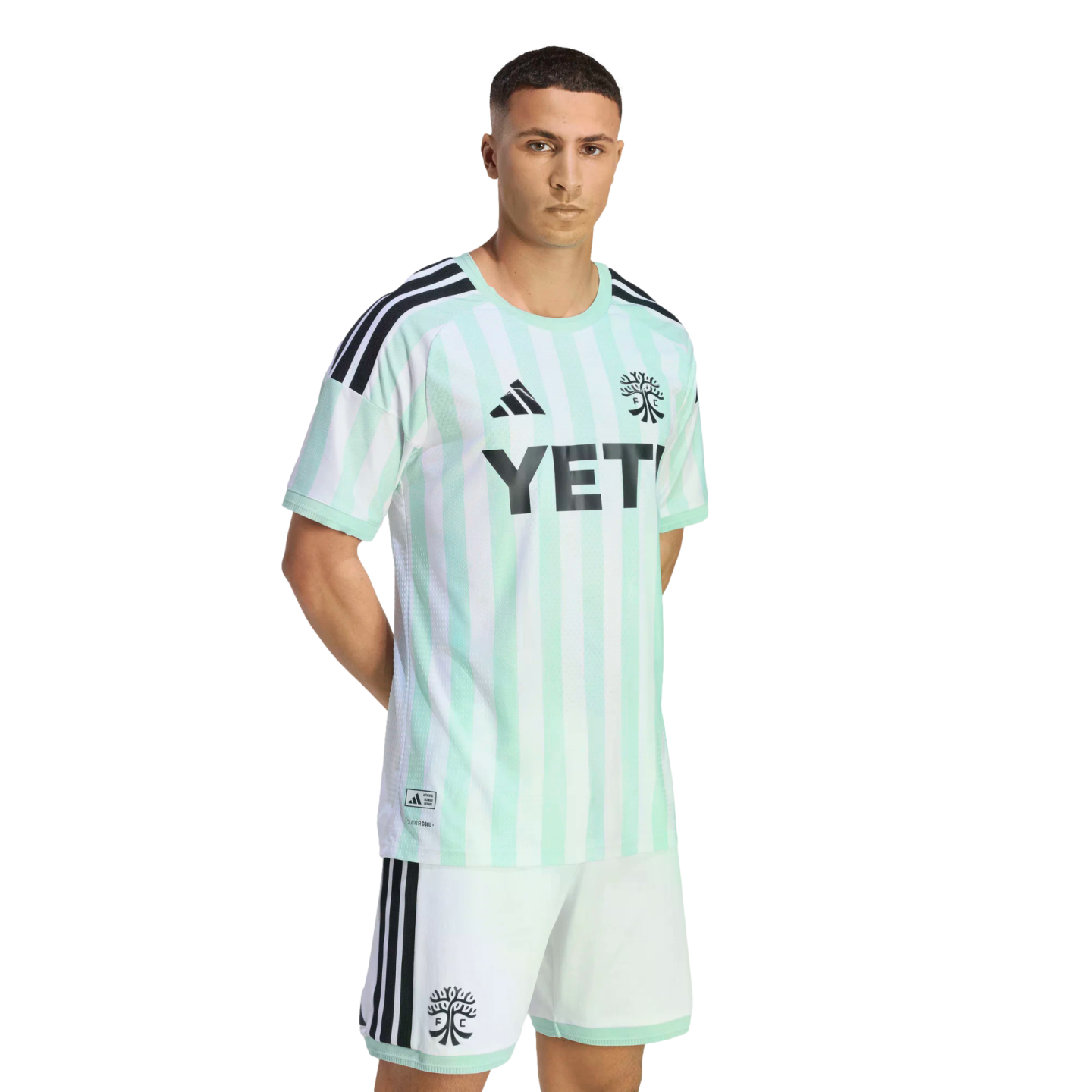 A man models a light green and white striped Austin FC soccer kit with black Adidas details and a YETI logo. He wears matching white shorts against a black background.