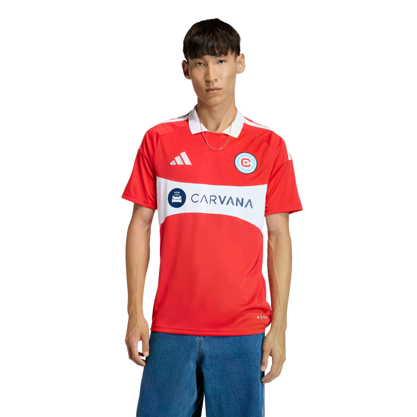A man models a red Chicago Fire soccer jersey featuring a white chest band with the "CARVANA" logo, paired with blue jeans against a black background.