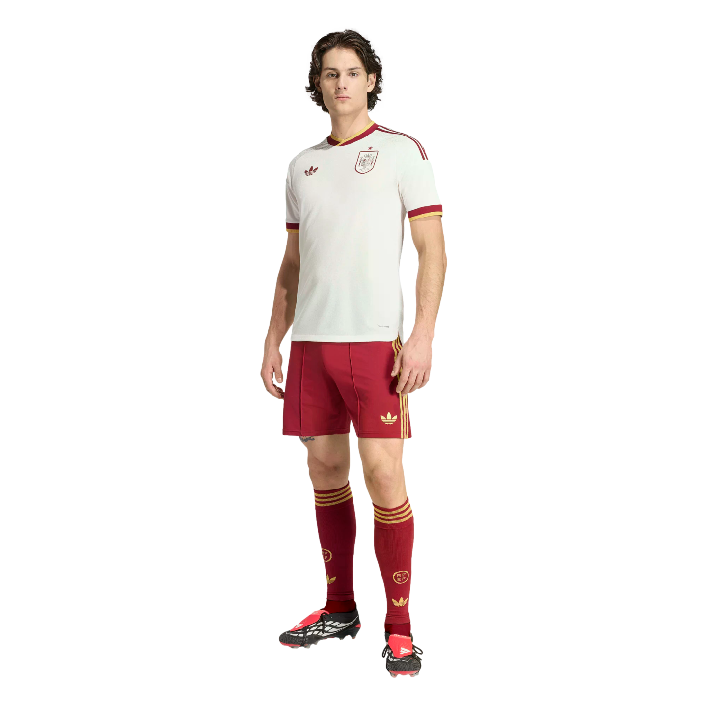 A man models a white and maroon Adidas Spain national football team kit against a black background.