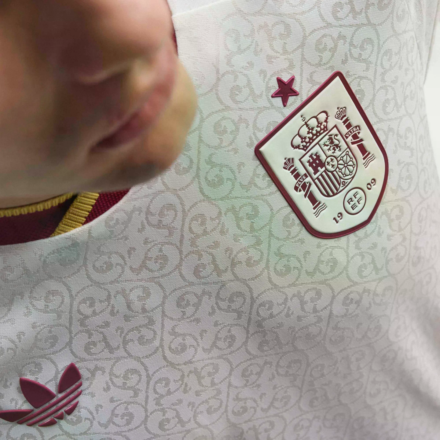 Close-up of a white Spain national team soccer jersey with a subtle ornate pattern, featuring a maroon RFEF crest and star, and an Adidas trefoil logo.