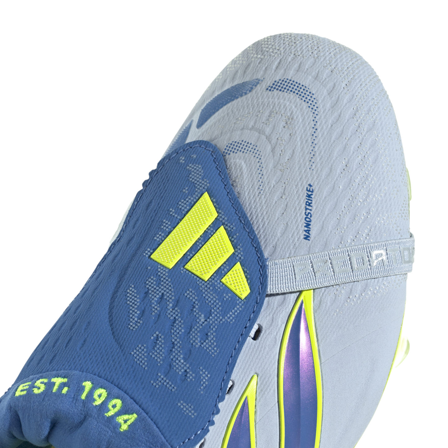 A close-up view of a light blue and blue Adidas Predator soccer cleat. The shoe features a blue fold-over tongue with neon yellow Adidas stripes and the text "EST. 1994" at its base. The light blue textured upper includes the text "NANOSTRIKE+" and a stra