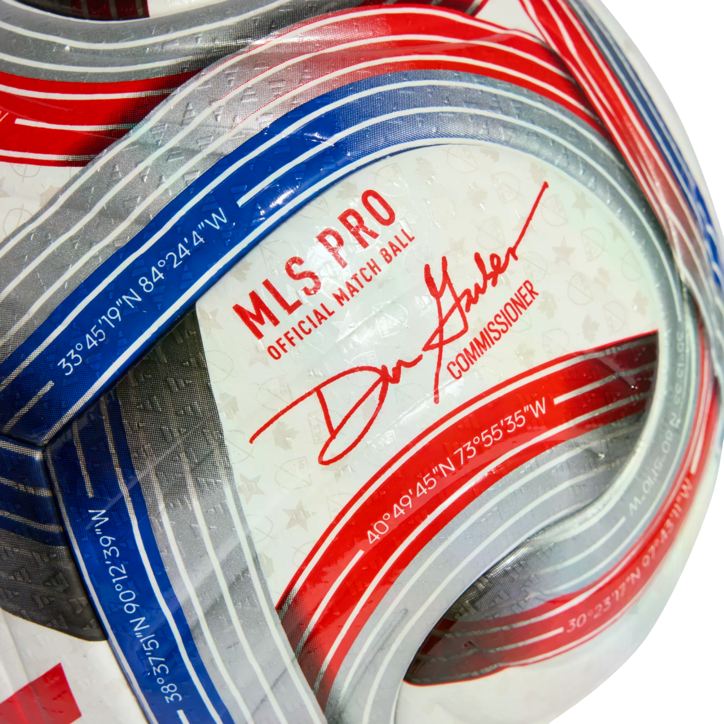 Close-up of a white MLS PRO soccer ball with red and blue stripes, the commissioner’s signature, and geographic coordinates printed on the surface.