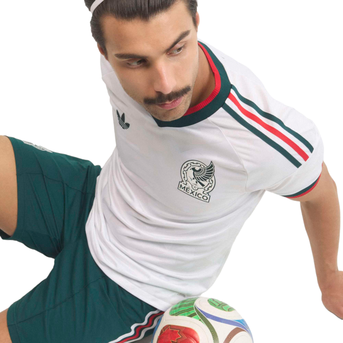 A man with a mustache wearing a white Mexico national soccer team jersey and green shorts sits with a soccer ball at his feet against a black background.