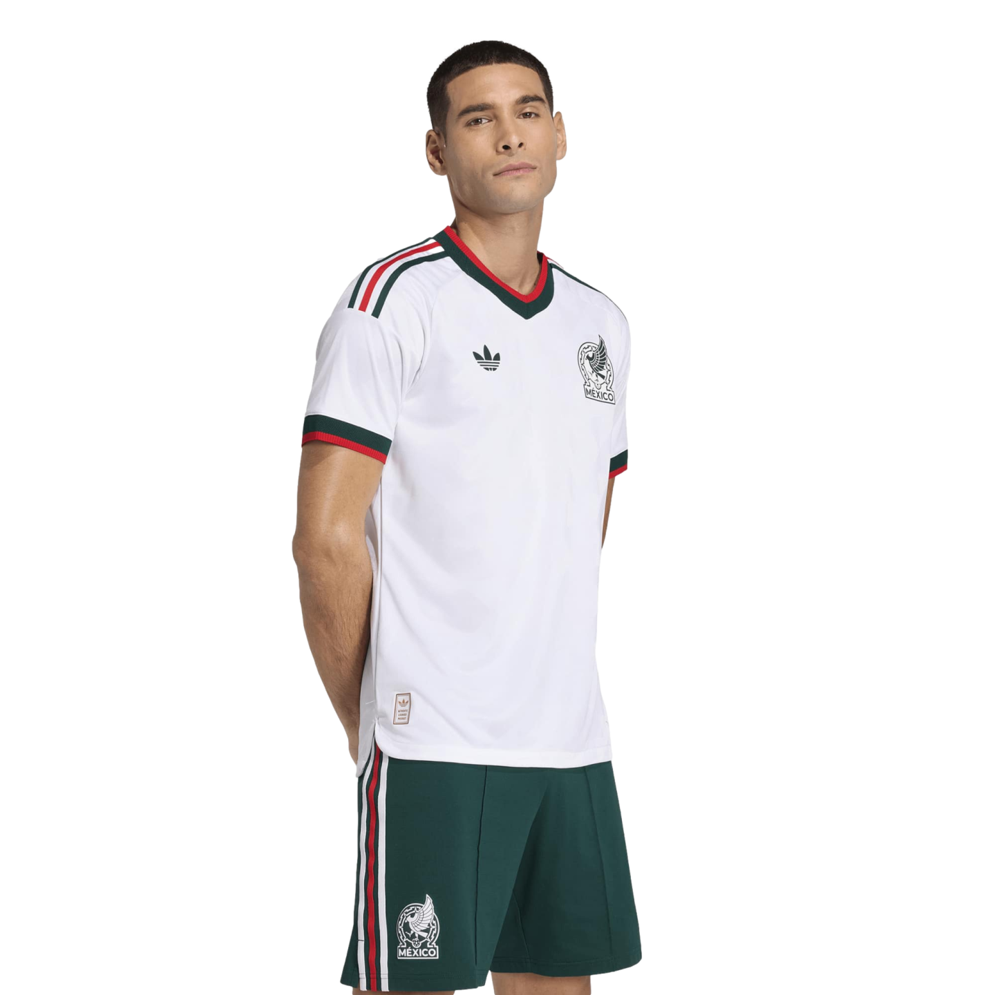 Man modeling a white Mexico national football team jersey and green shorts with red and white accents against a black background.