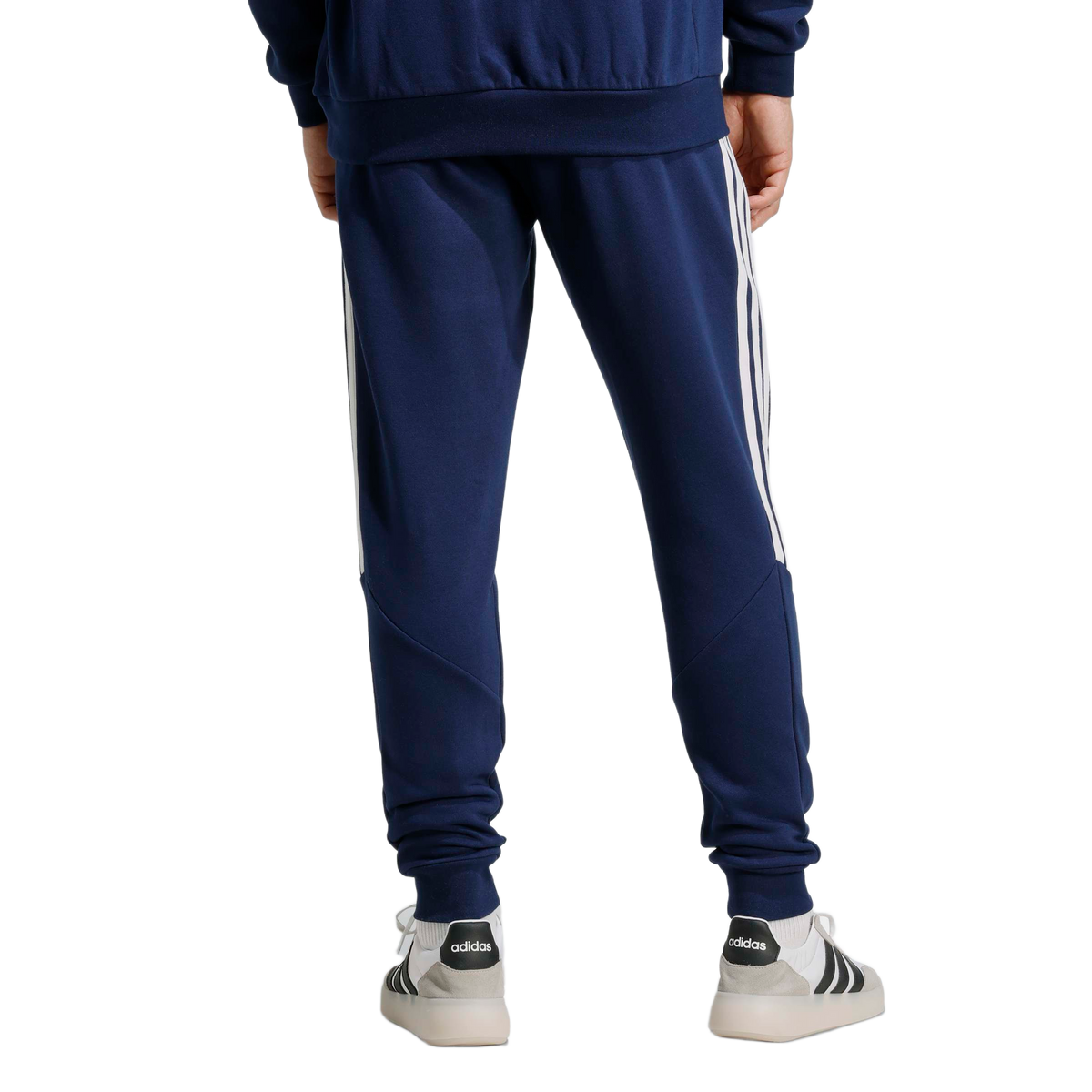 Rear view of a person wearing navy Adidas track pants with white side stripes and white sneakers against a light gray background.