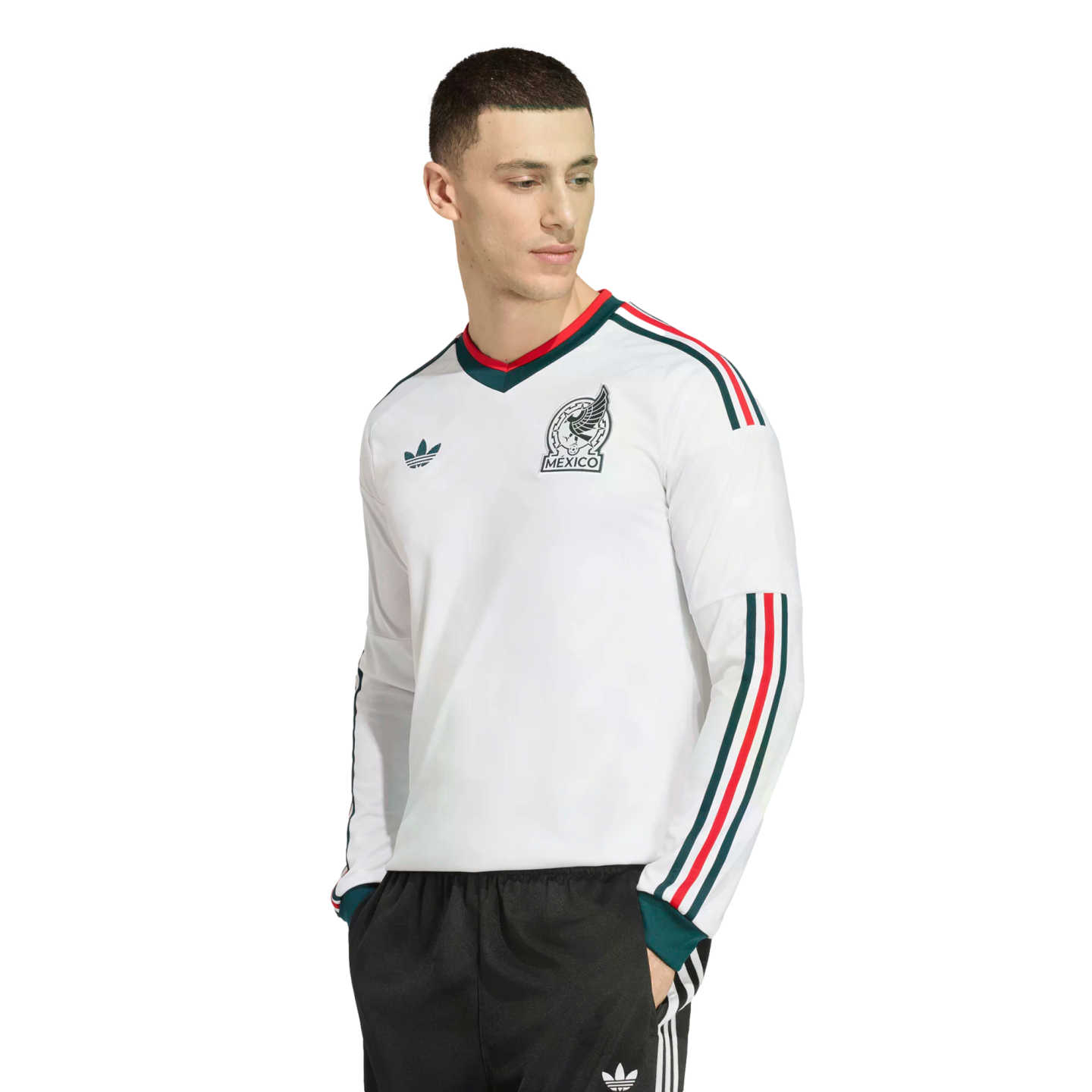 A man wearing a white long-sleeved Adidas Mexico soccer jersey with green and red trim, paired with black tracksuit pants, against a black background.