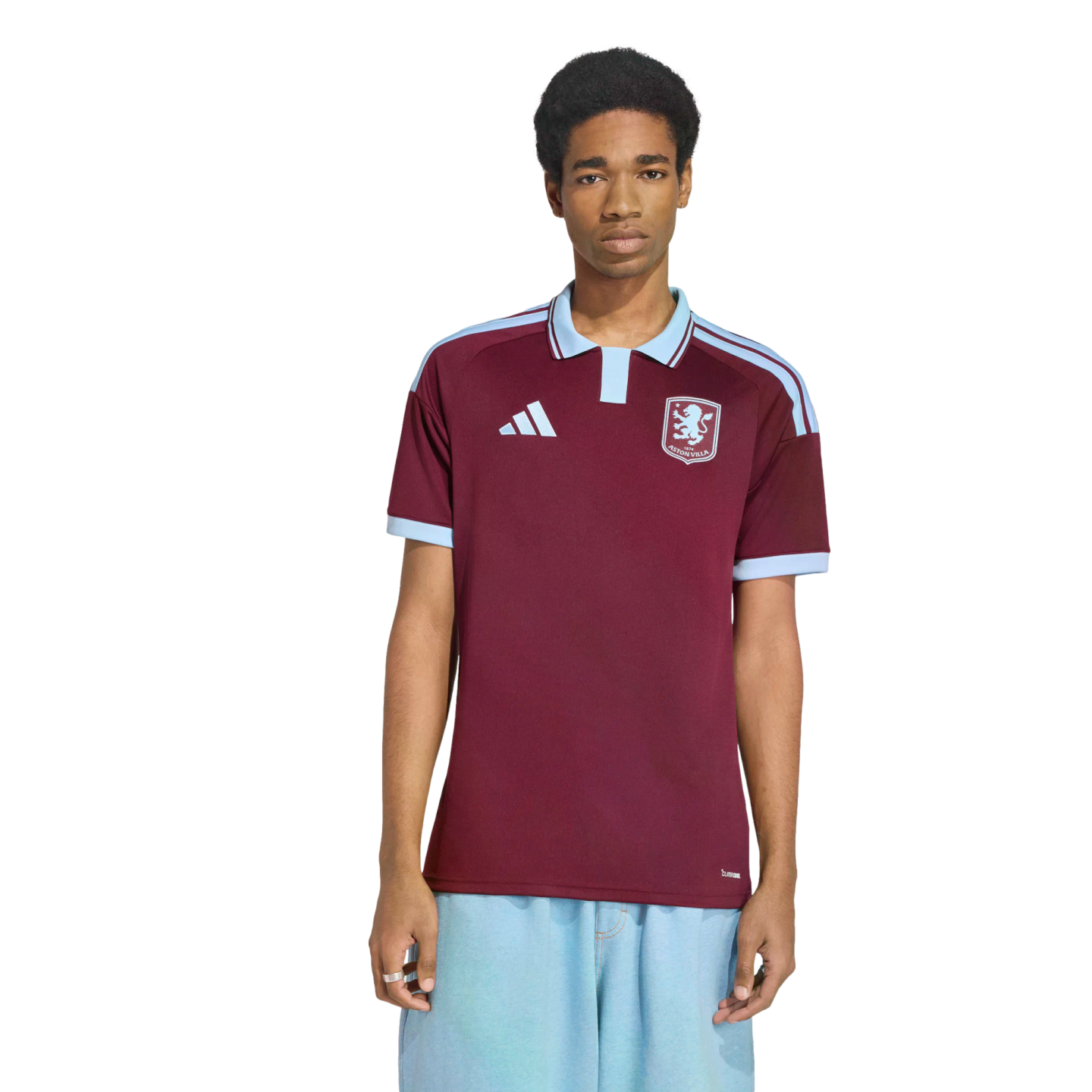 A man models a maroon and light-blue Adidas soccer jersey with a polo collar and a lion crest on the chest, standing against a white background.