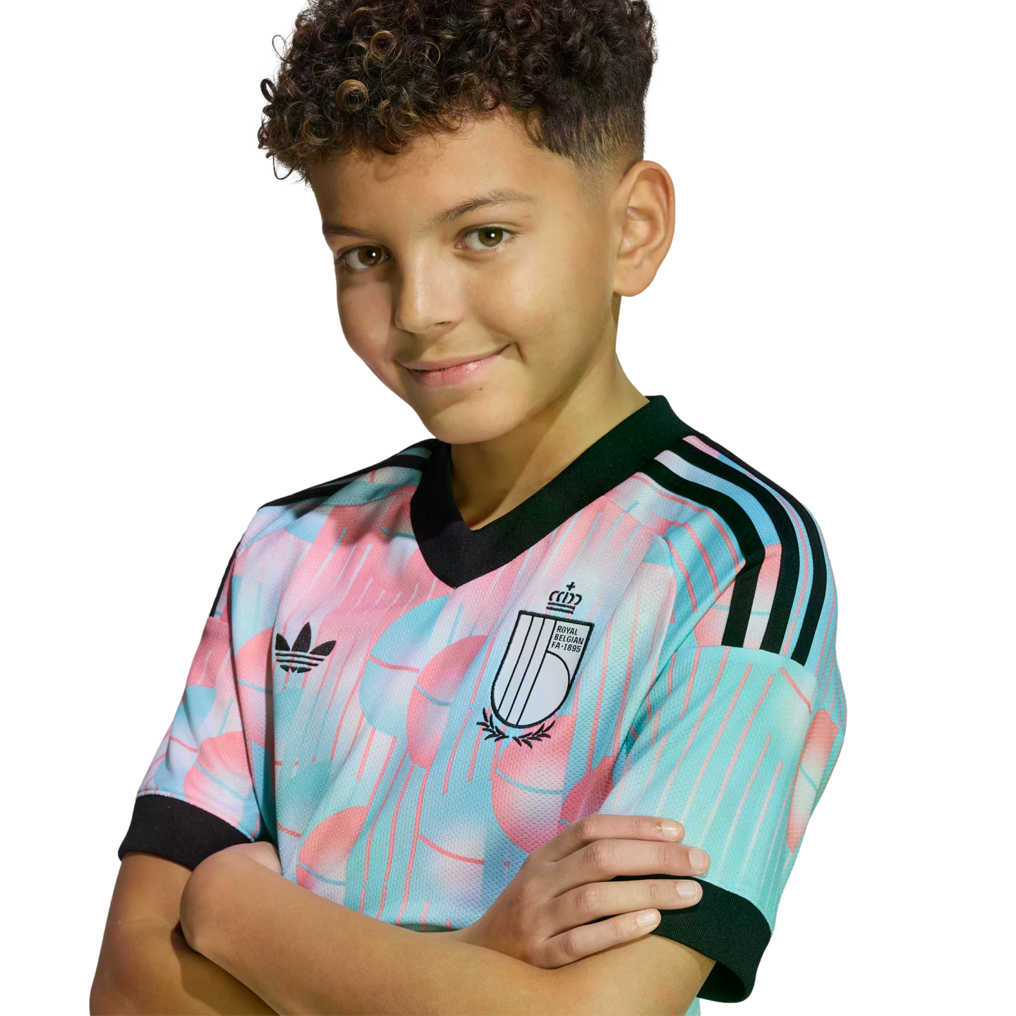 A smiling boy with curly hair poses with arms crossed, wearing a colorful pastel Belgian national football team jersey against a black background.