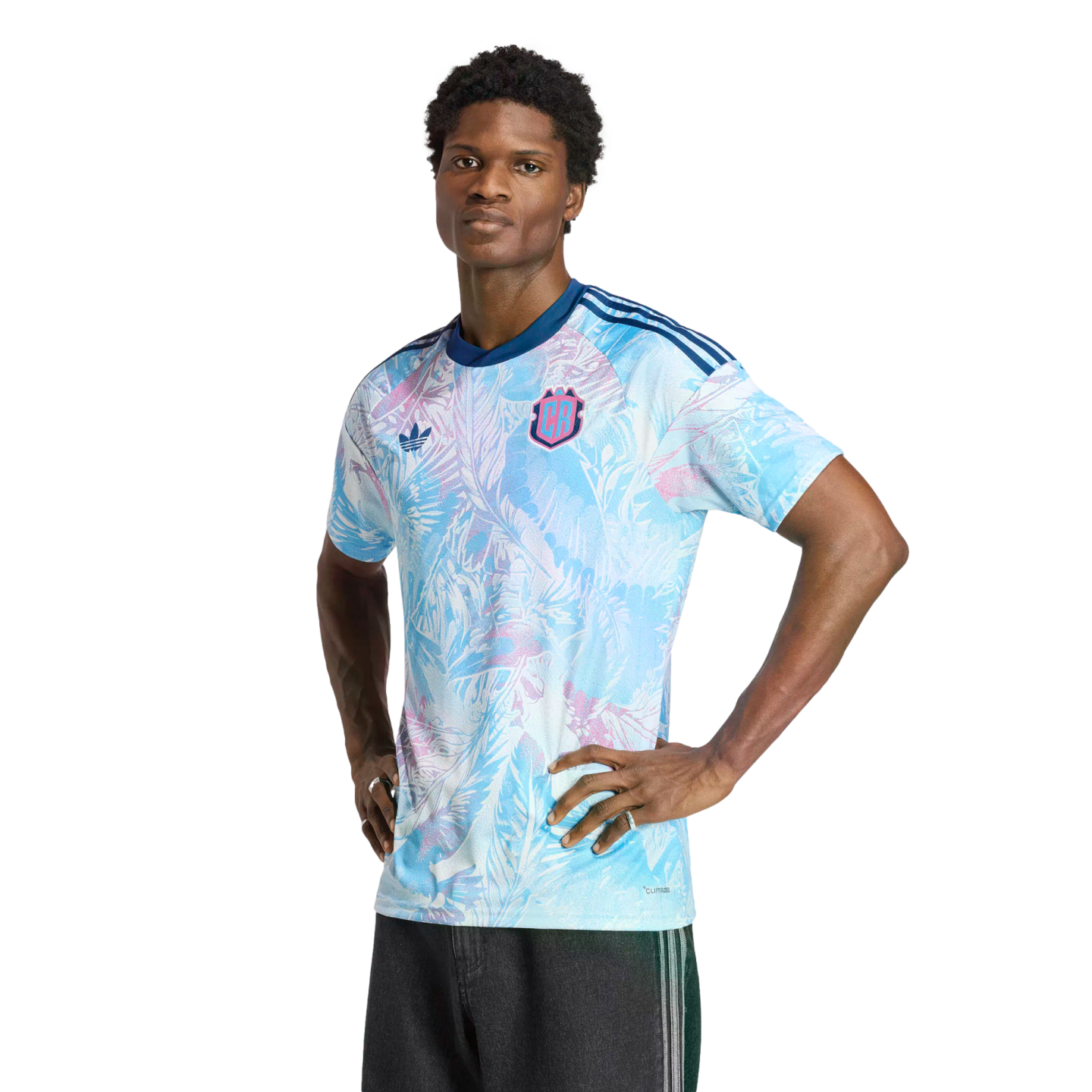A man models a light blue and pink floral-patterned Adidas jersey with a 'CR' crest, standing with his hands on his hips against a solid black background.