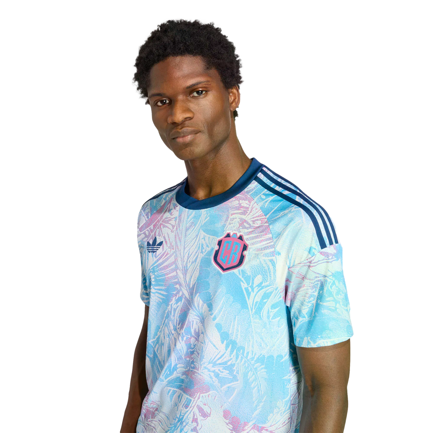 A man models a light blue and pink tropical-print Adidas jersey with a "CB" crest against a black background.