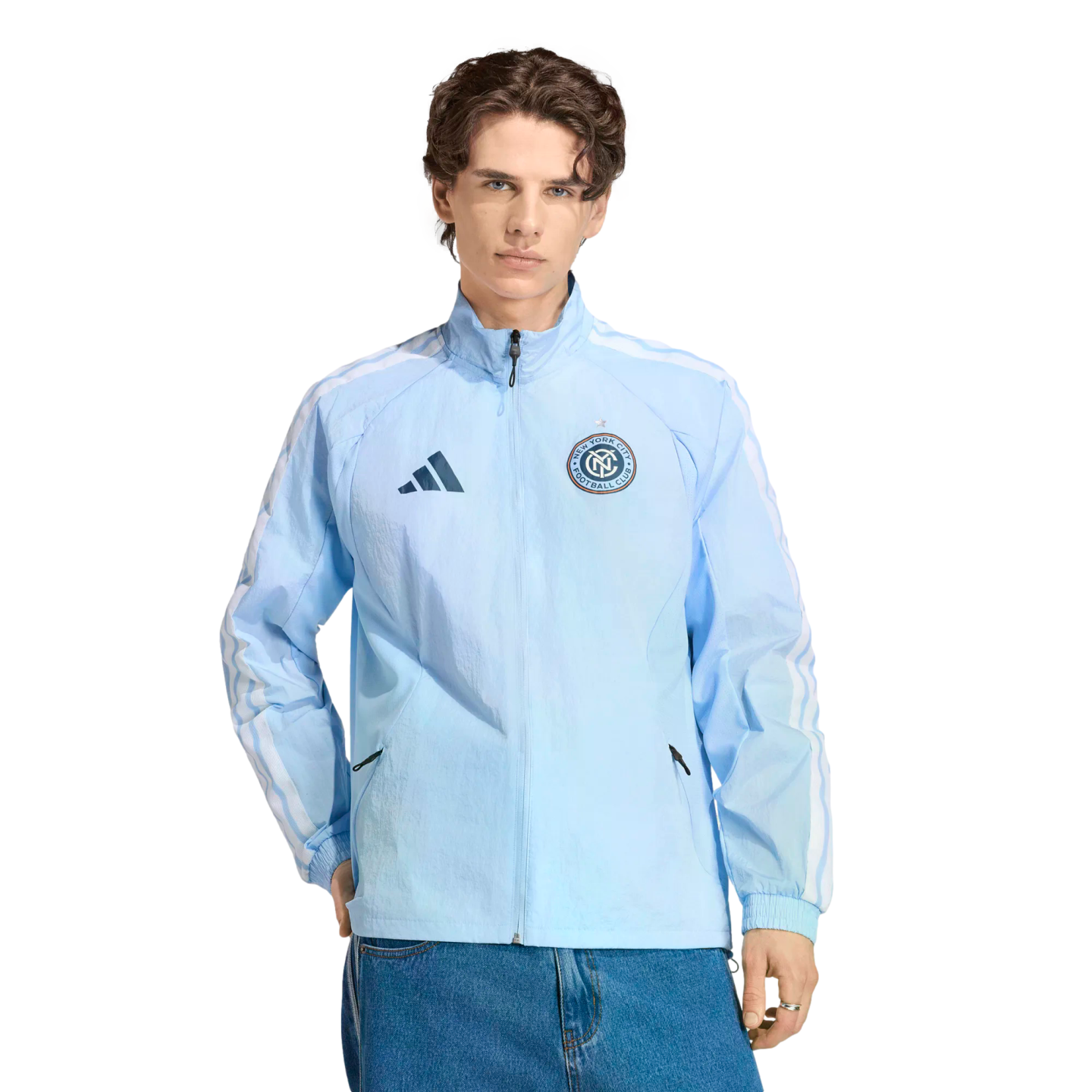 A man wearing a light blue Adidas New York City FC windbreaker and blue jeans against a black background.