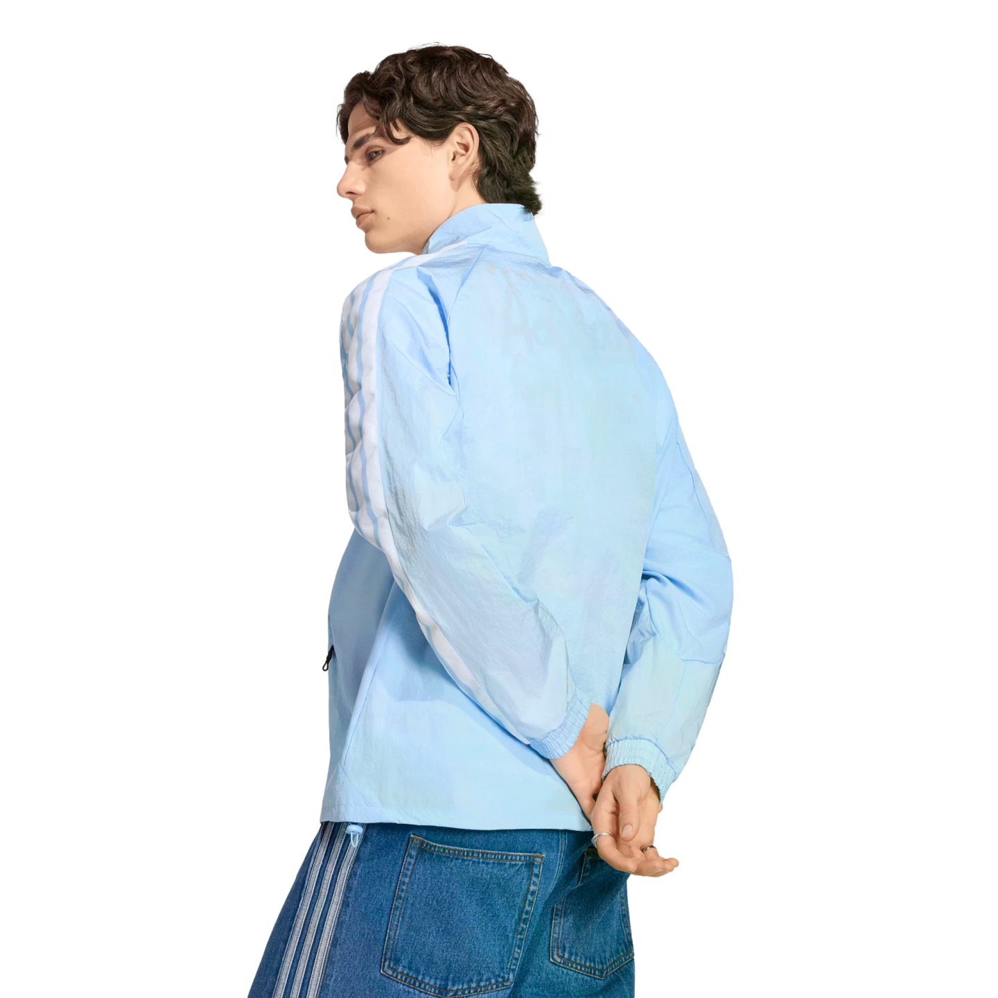 A man in a light blue zip-up track jacket and denim jeans looks back over his shoulder against a black background.
