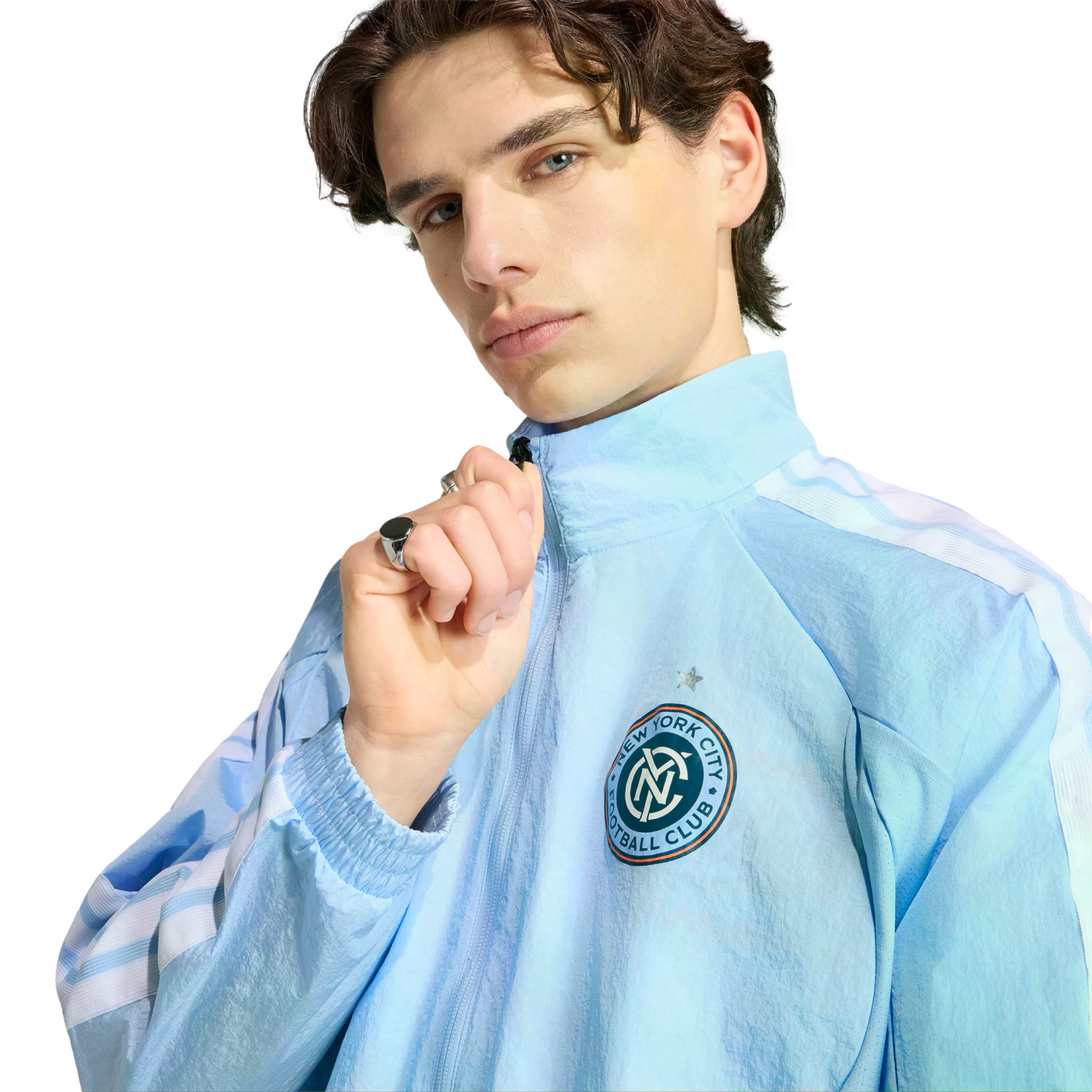 A man with dark wavy hair wears a light blue New York City Football Club zip-up jacket, posing against a black background.