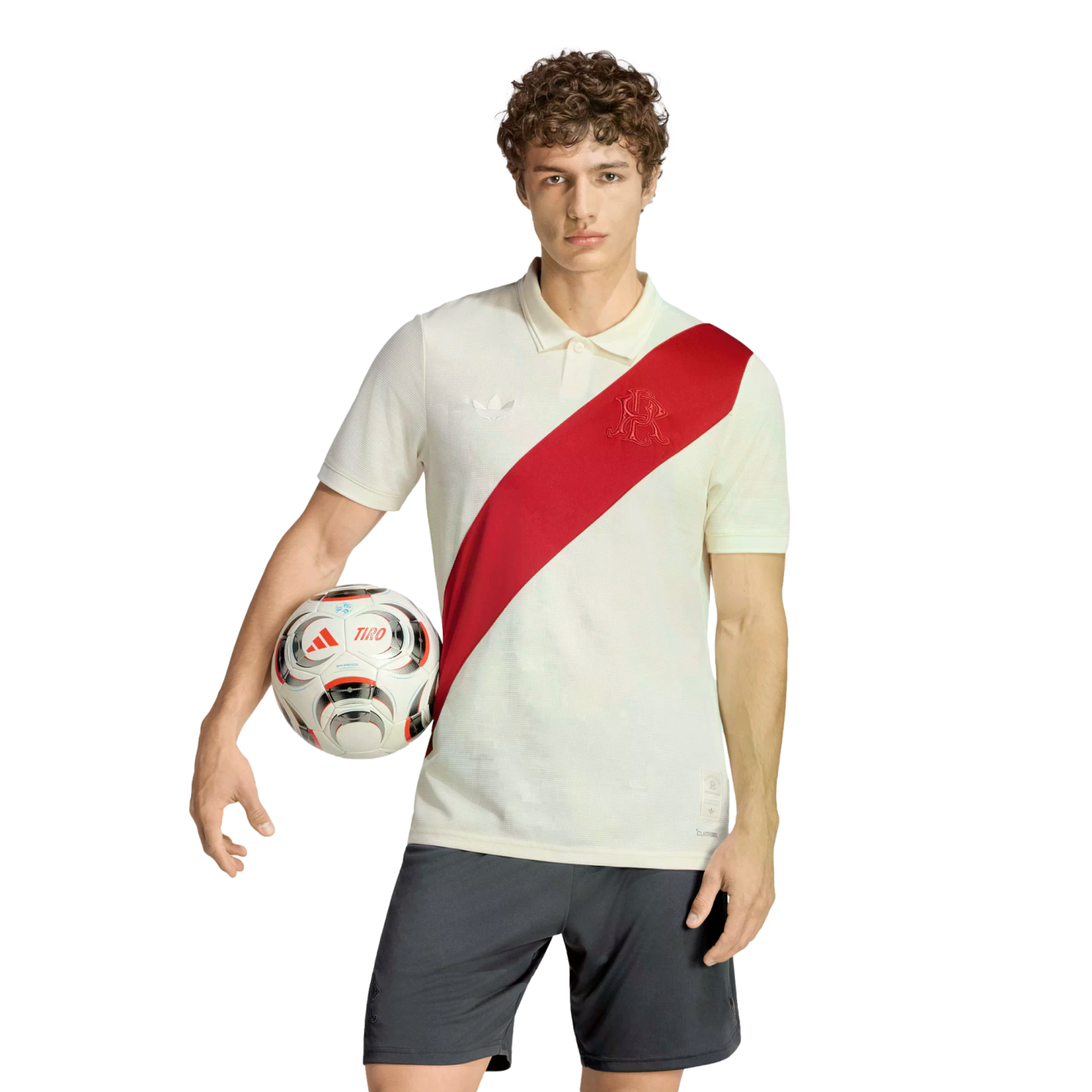 A man with curly hair stands against a white background, wearing an off-white polo with a red diagonal sash and dark shorts, holding a soccer ball under his arm.