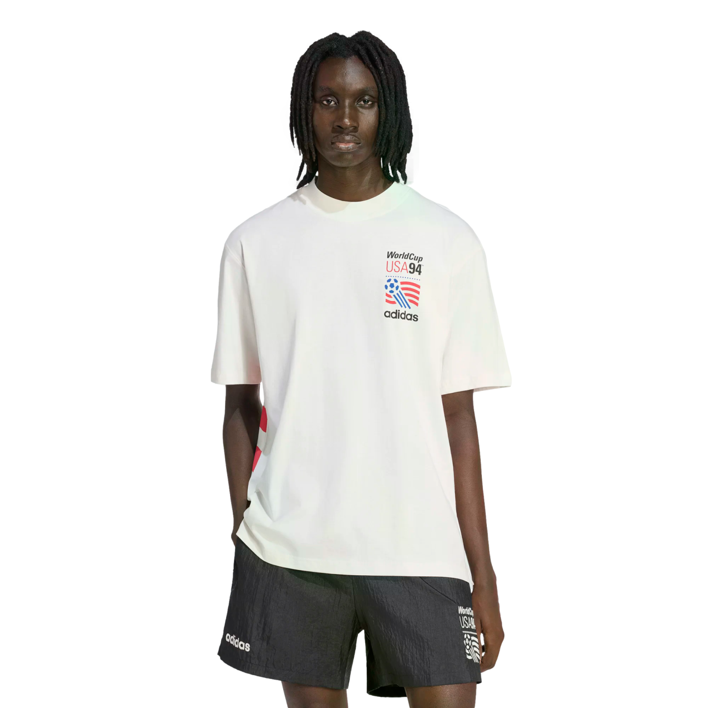 A Black man with locs wearing a white Adidas WorldCup USA 94 graphic T-shirt and matching black shorts against a white background.